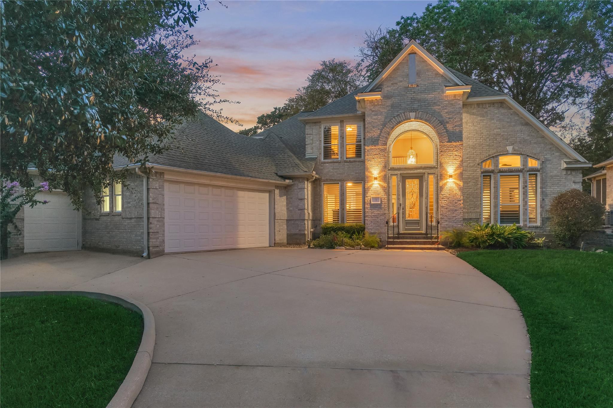 Stone house at dusk with a tall arched entry, glowing windows, and a broad curved driveway.
