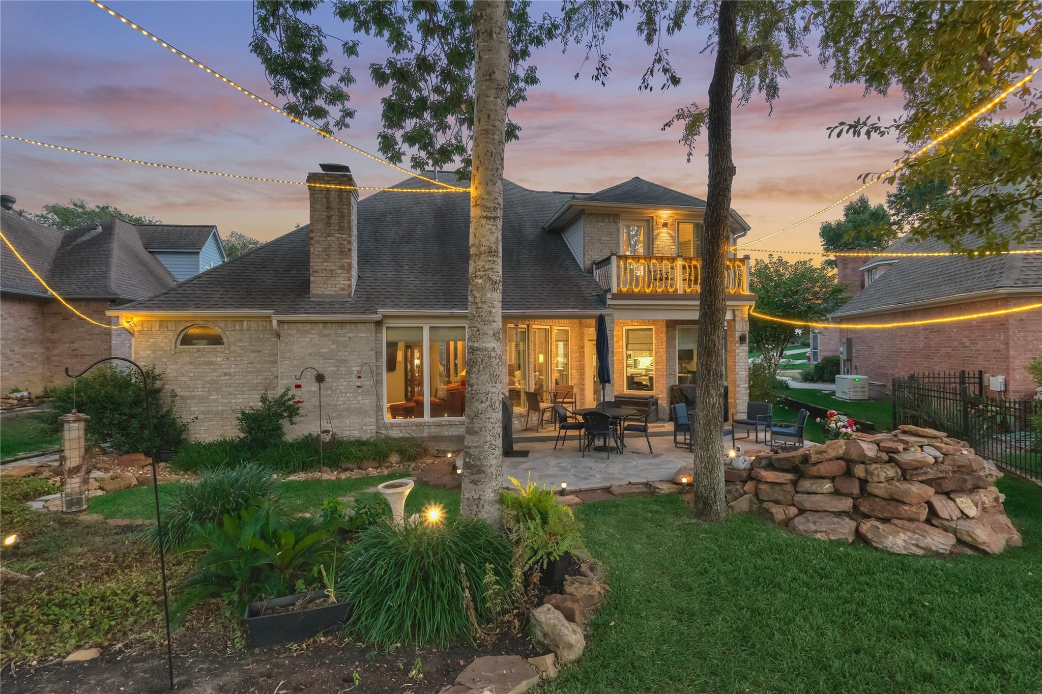Brick home’s backyard patio at dusk, lit by string lights and surrounded by landscaped gardens.