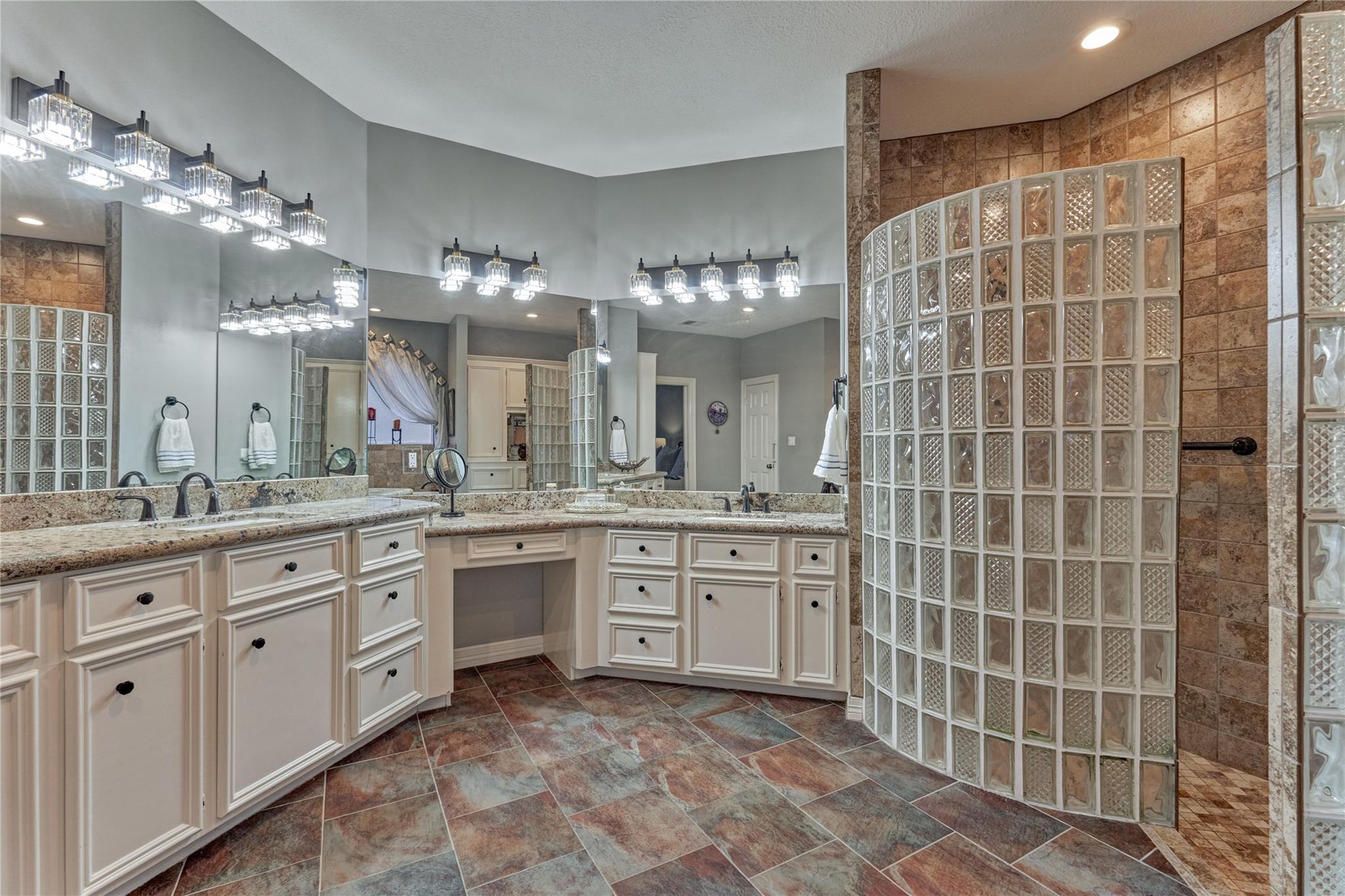 Primary bathroom with a curved glass-block shower, mirrored walls, white cabinetry, and multicolored tile flooring.