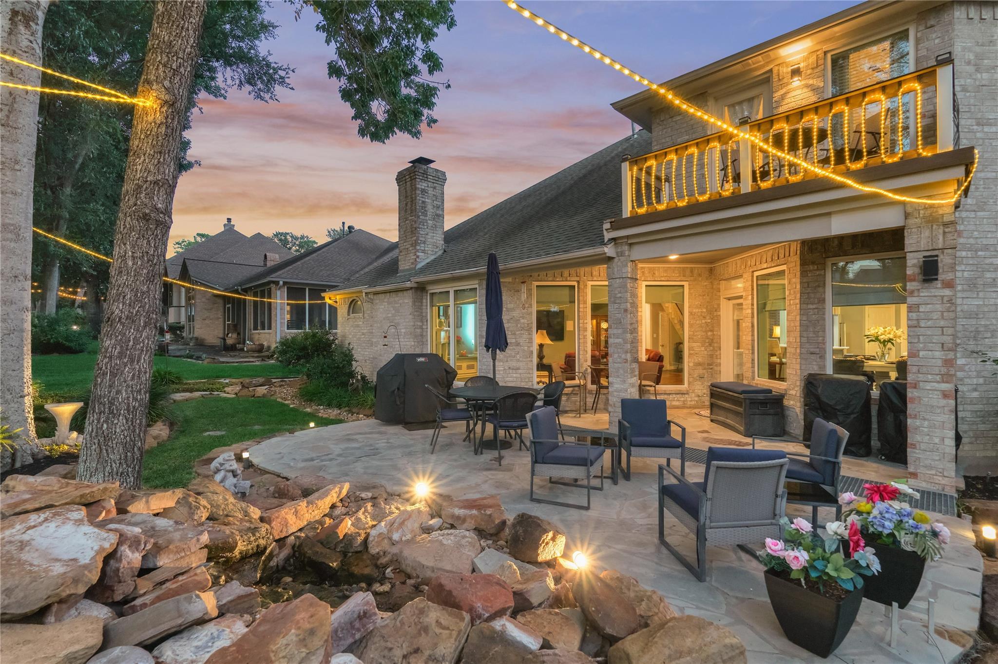 Stone home’s backyard patio at dusk, with balcony string lights, outdoor seating, and a rock water feature.