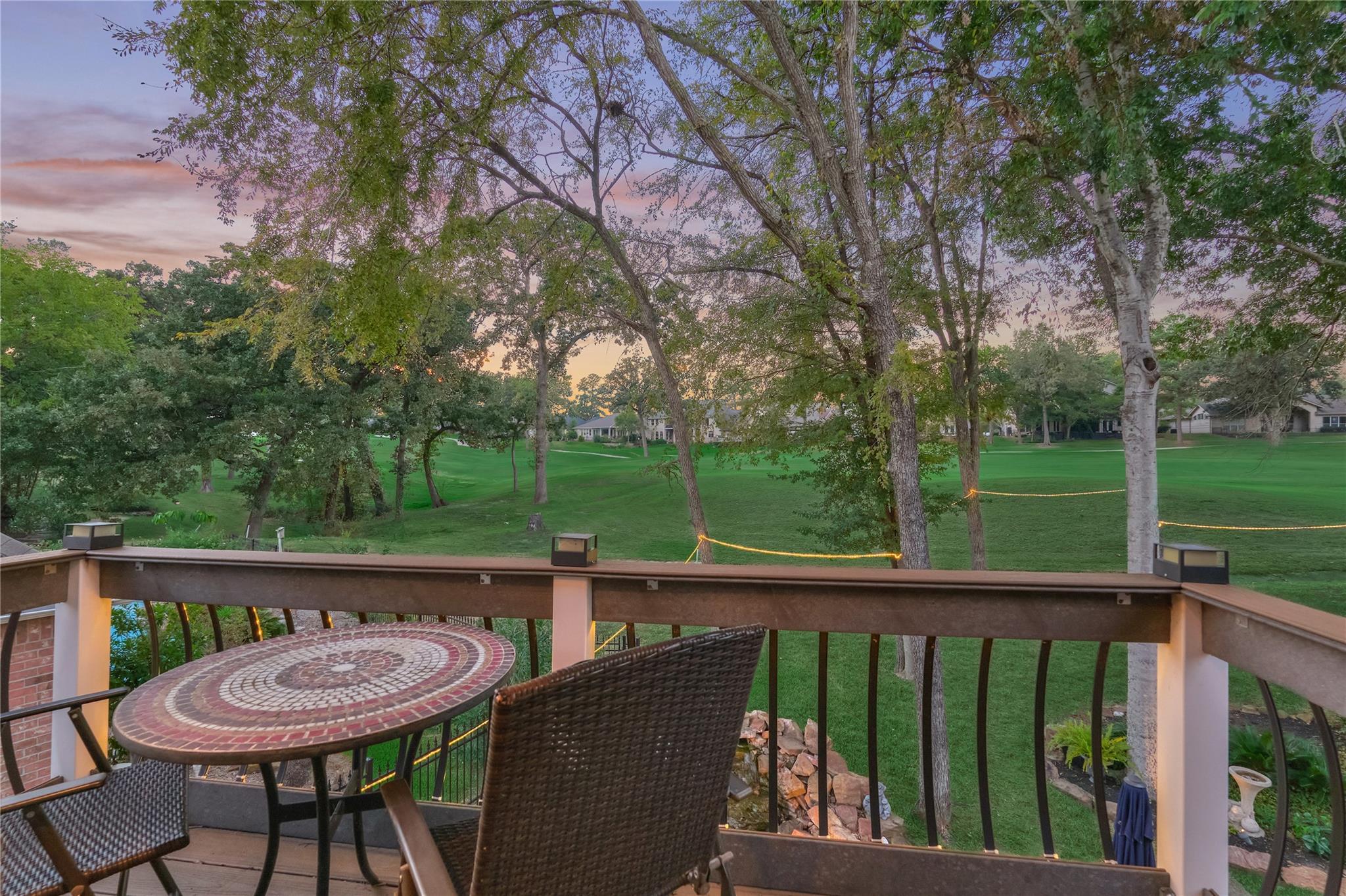 Furnished deck with a mosaic table overlooks a tree-lined green lawn at sunset.