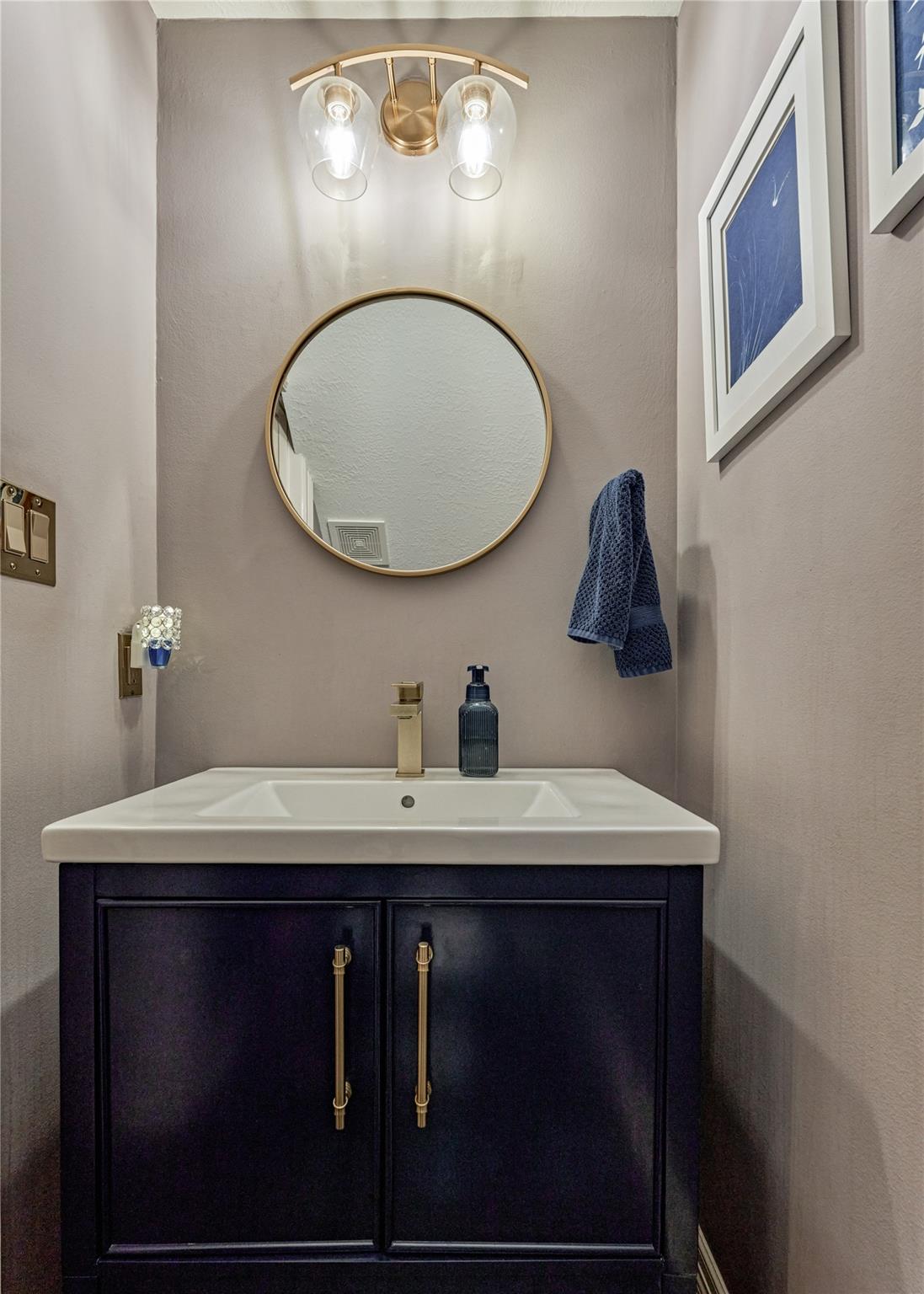 Bathroom vanity with a white sink, navy cabinet, brass fixtures, and round gold-framed mirror.