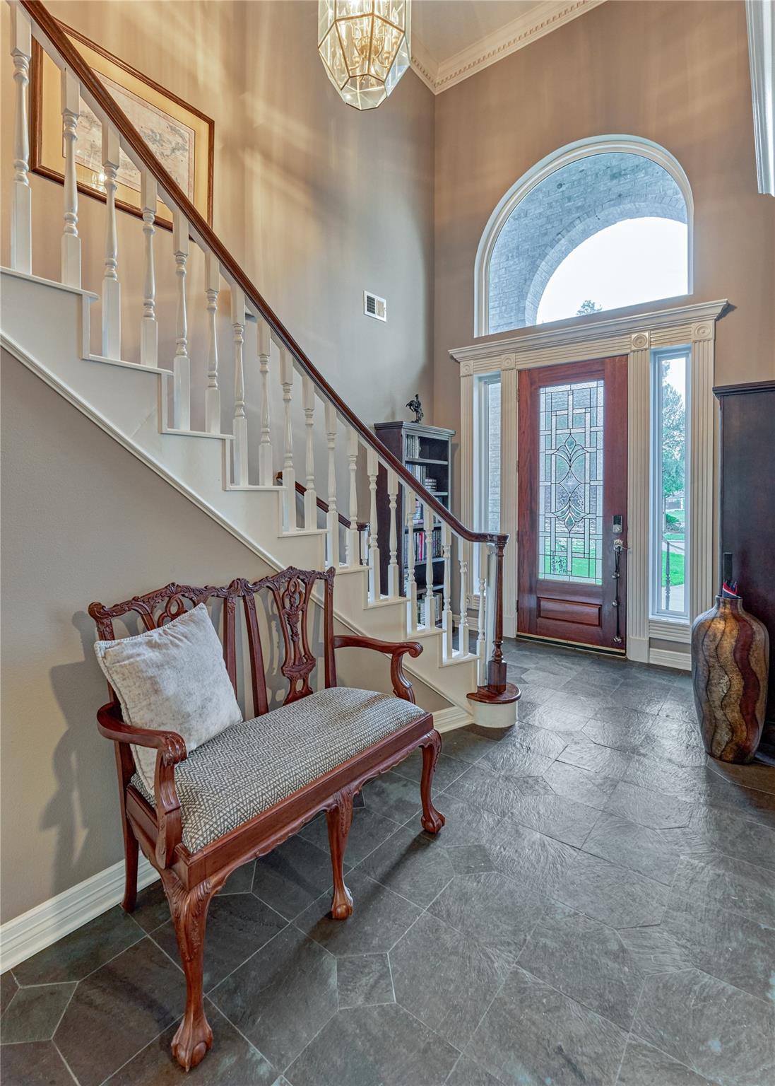 Foyer with a sweeping staircase, carved wood bench, and stained-glass front door beneath an arched window.