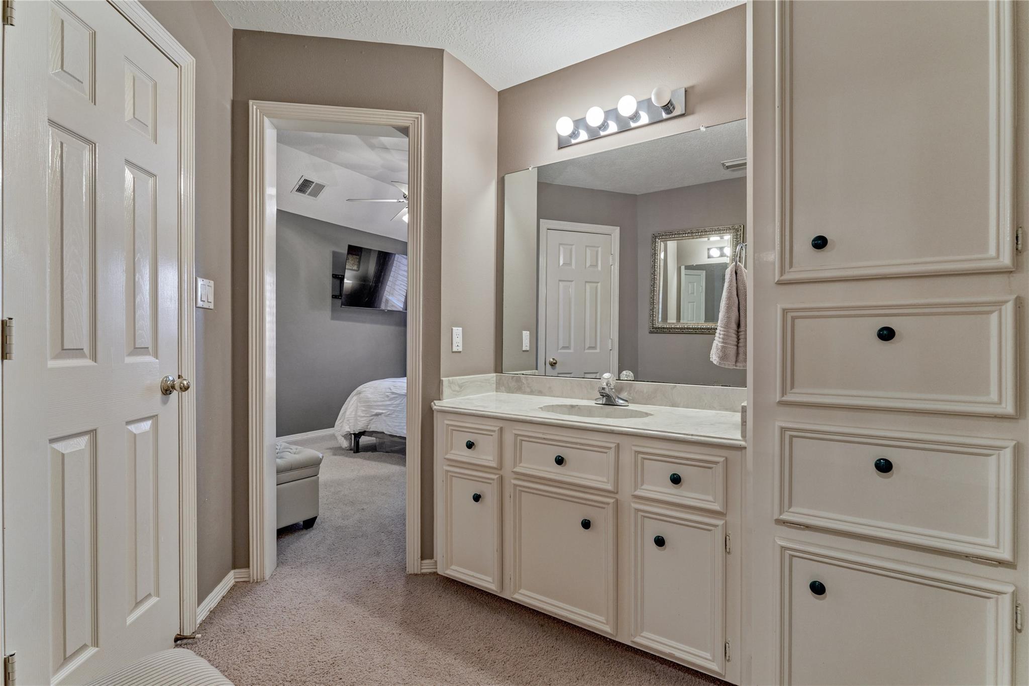 Bathroom vanity with a large mirror and cream cabinetry, opening onto a carpeted bedroom.