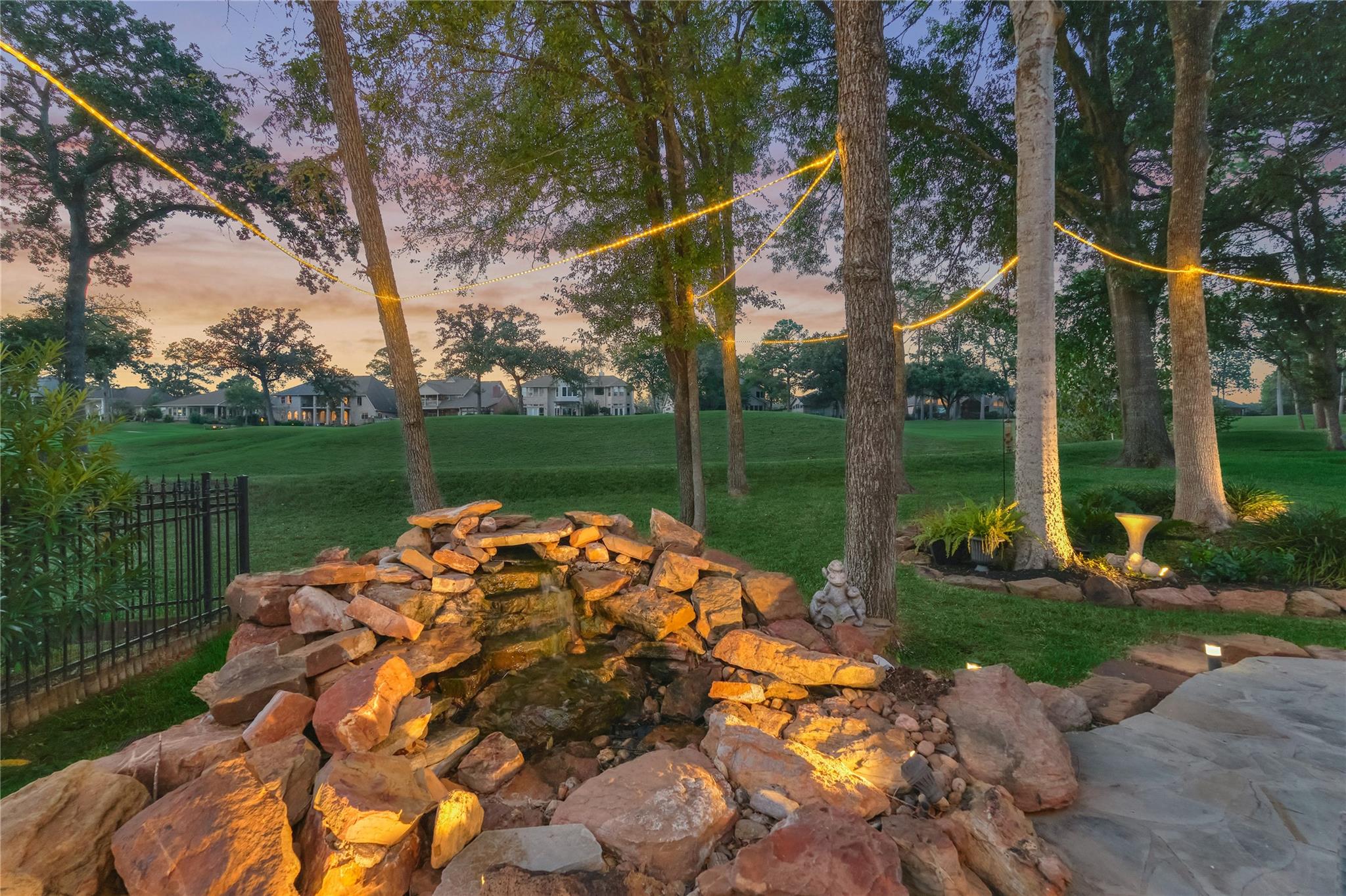Rock waterfall and pond in a landscaped yard beneath mature trees and glowing string lights at dusk