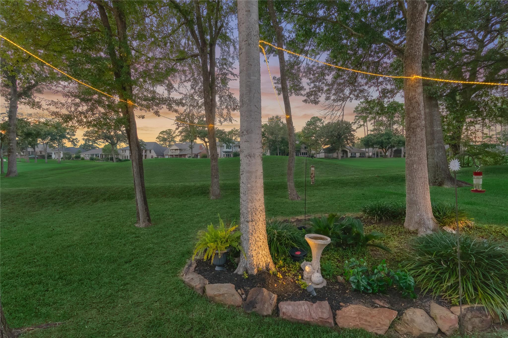 Tree-lined lawn at dusk with overhead string lights and a stone-edged garden bed.