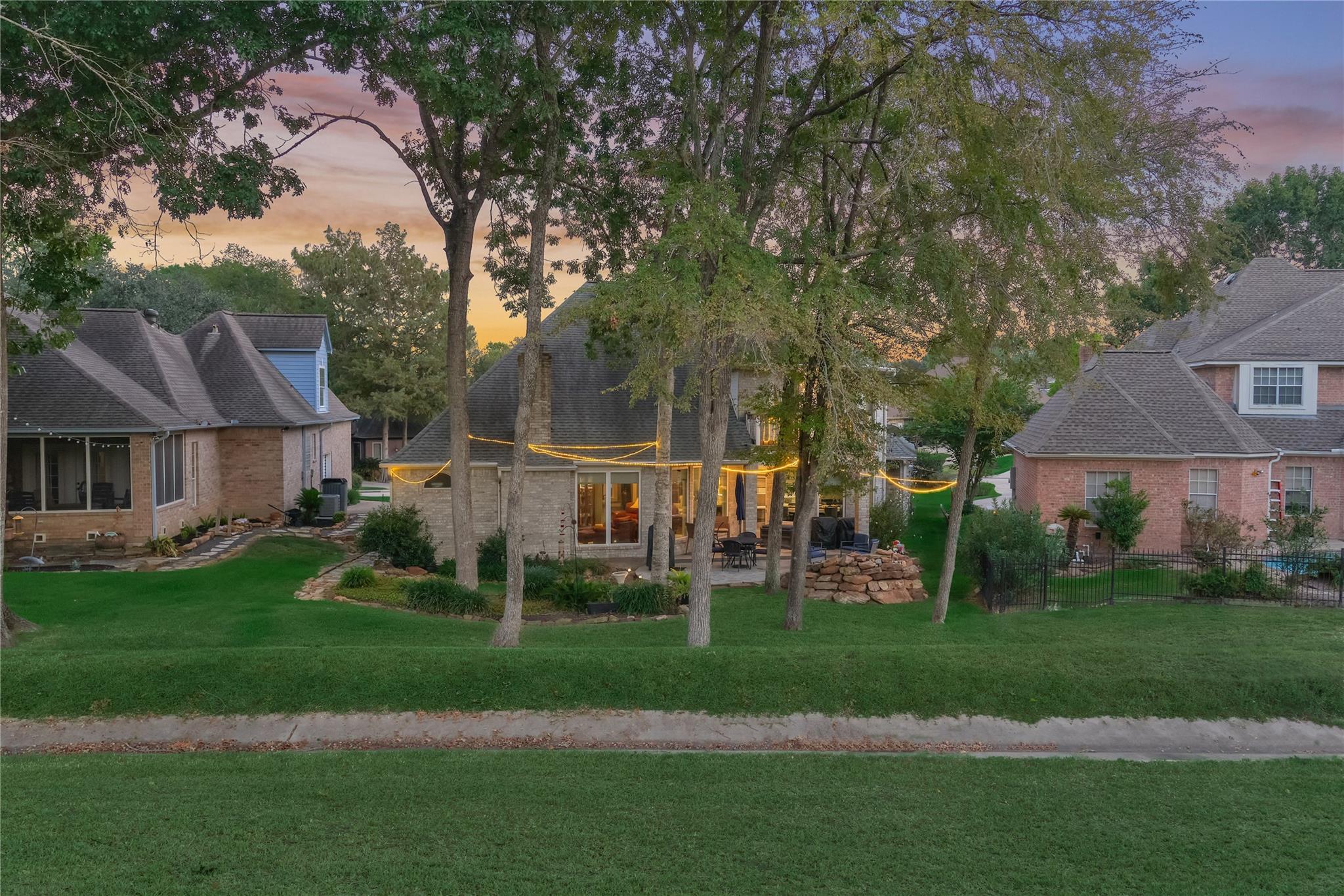 Two-story brick home at dusk with a tree-framed patio, glowing string lights, and a broad lawn.