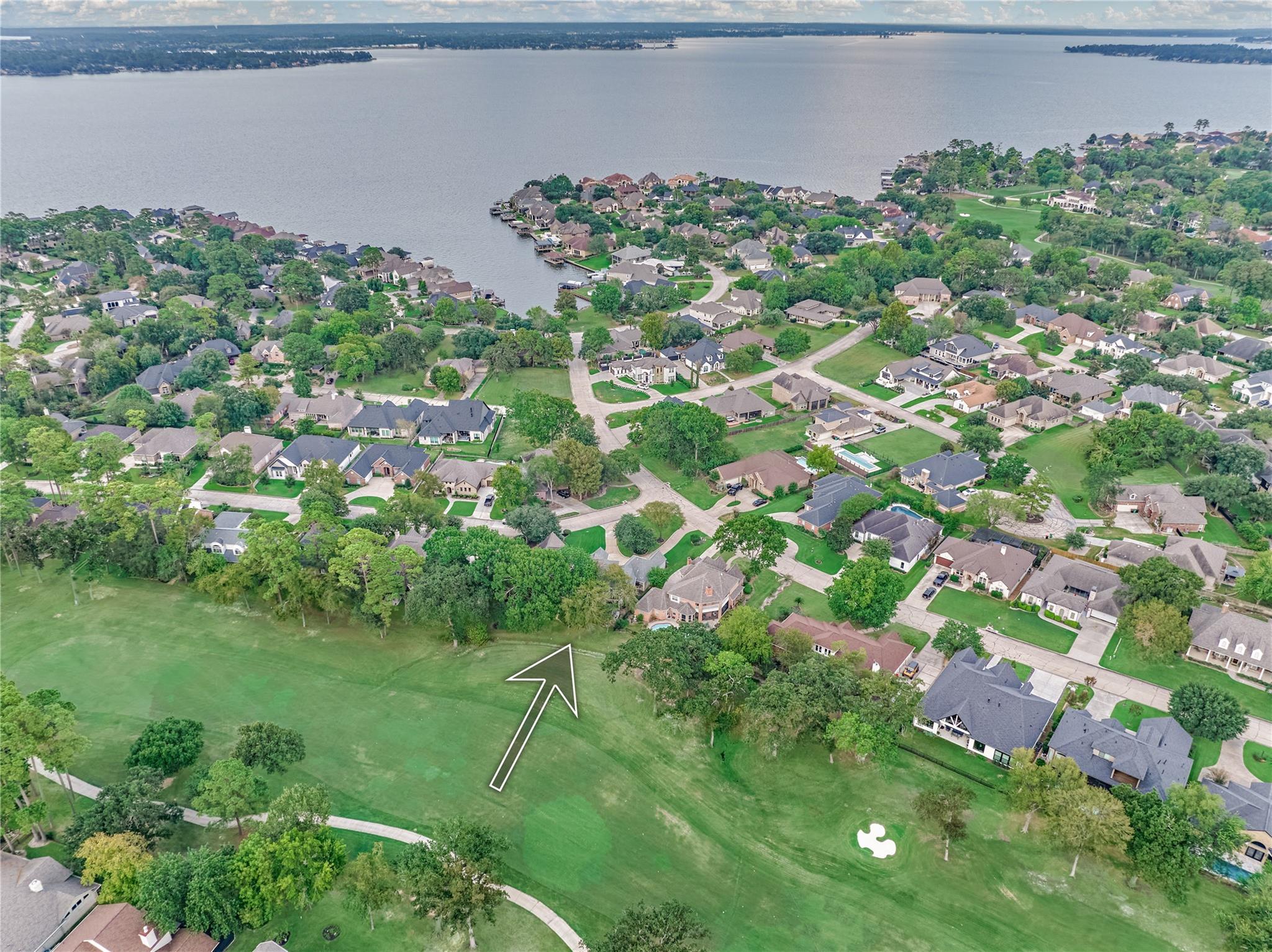 Aerial view of a lakeside neighborhood; an arrow marks a tree-lined lot beside a broad green space.