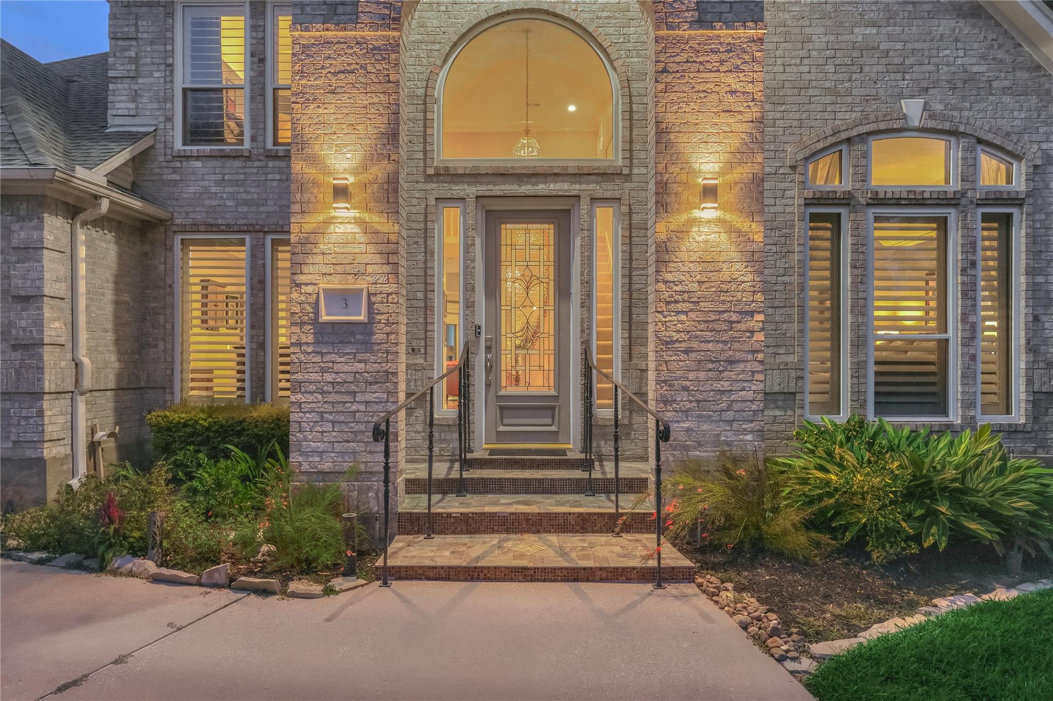 Gray brick home entrance with an arched window, decorative glass door, tiled steps, and landscaped beds.