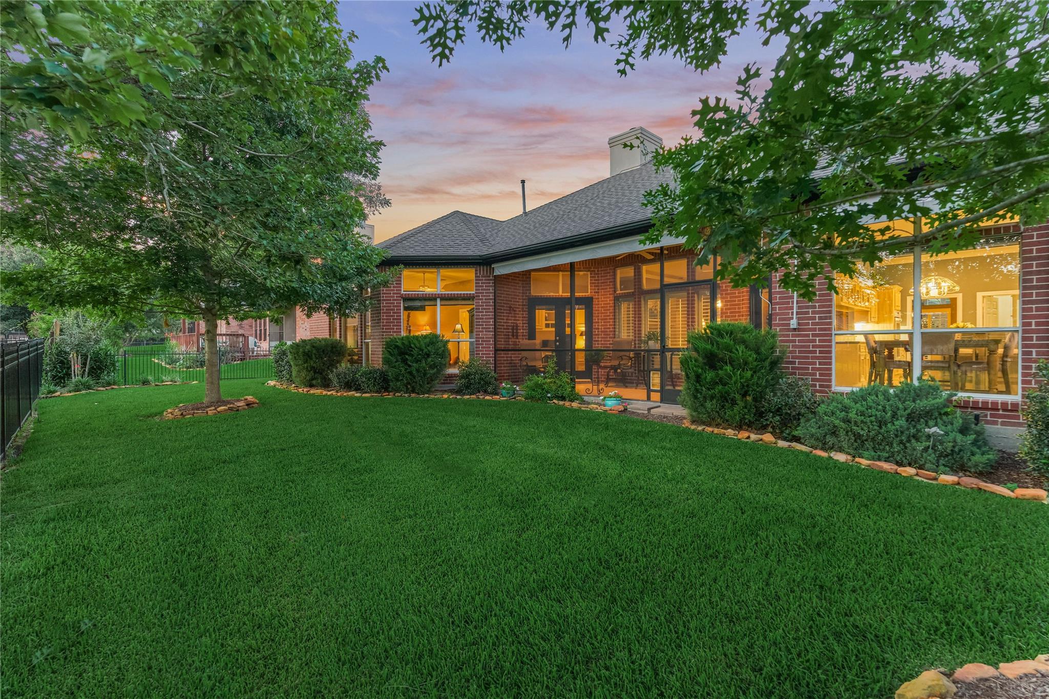 Red-brick home with glowing windows and a screened patio beyond a broad lawn at dusk.