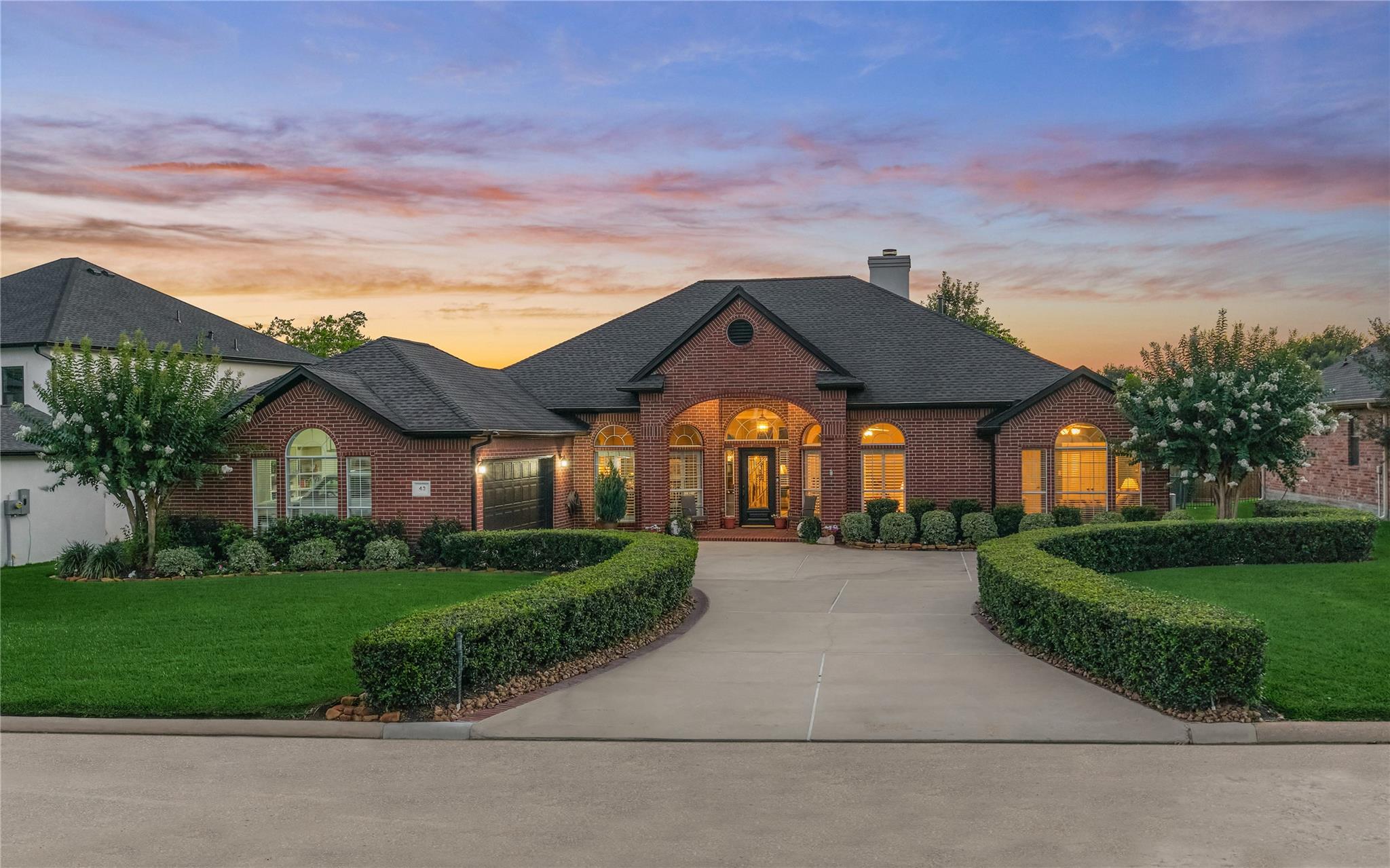 Red-brick home with arched windows, a lit entry, and a curved driveway at dusk