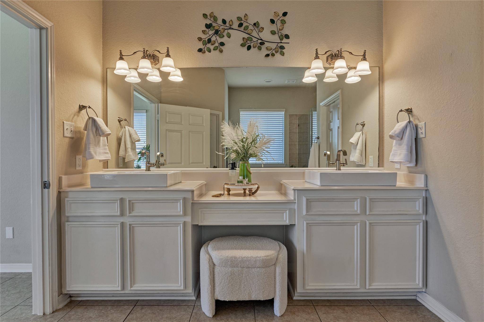 Bathroom double vanity with white vessel sinks, a large mirror, and a cushioned stool.