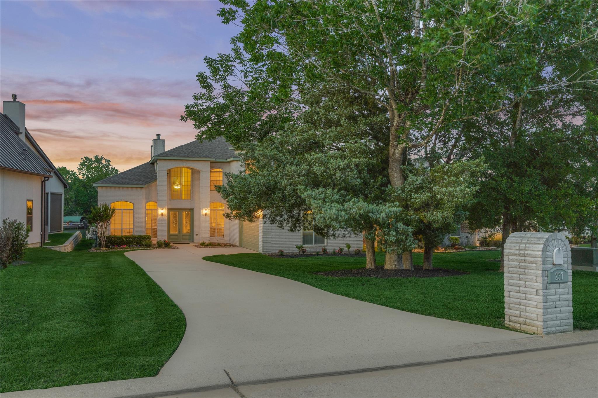 Two-story brick house with warmly lit windows, a curved driveway, mature trees, and a stone mailbox.