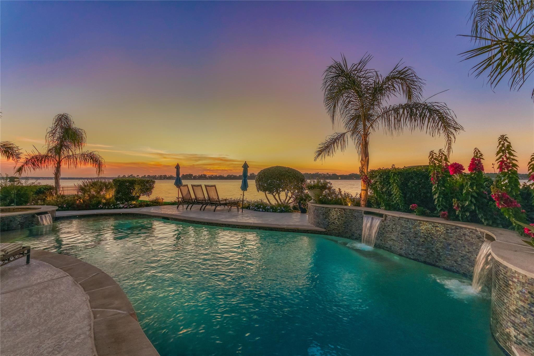 Curved swimming pool with stone waterfalls overlooks the water at sunset, framed by palms and flowering shrubs.