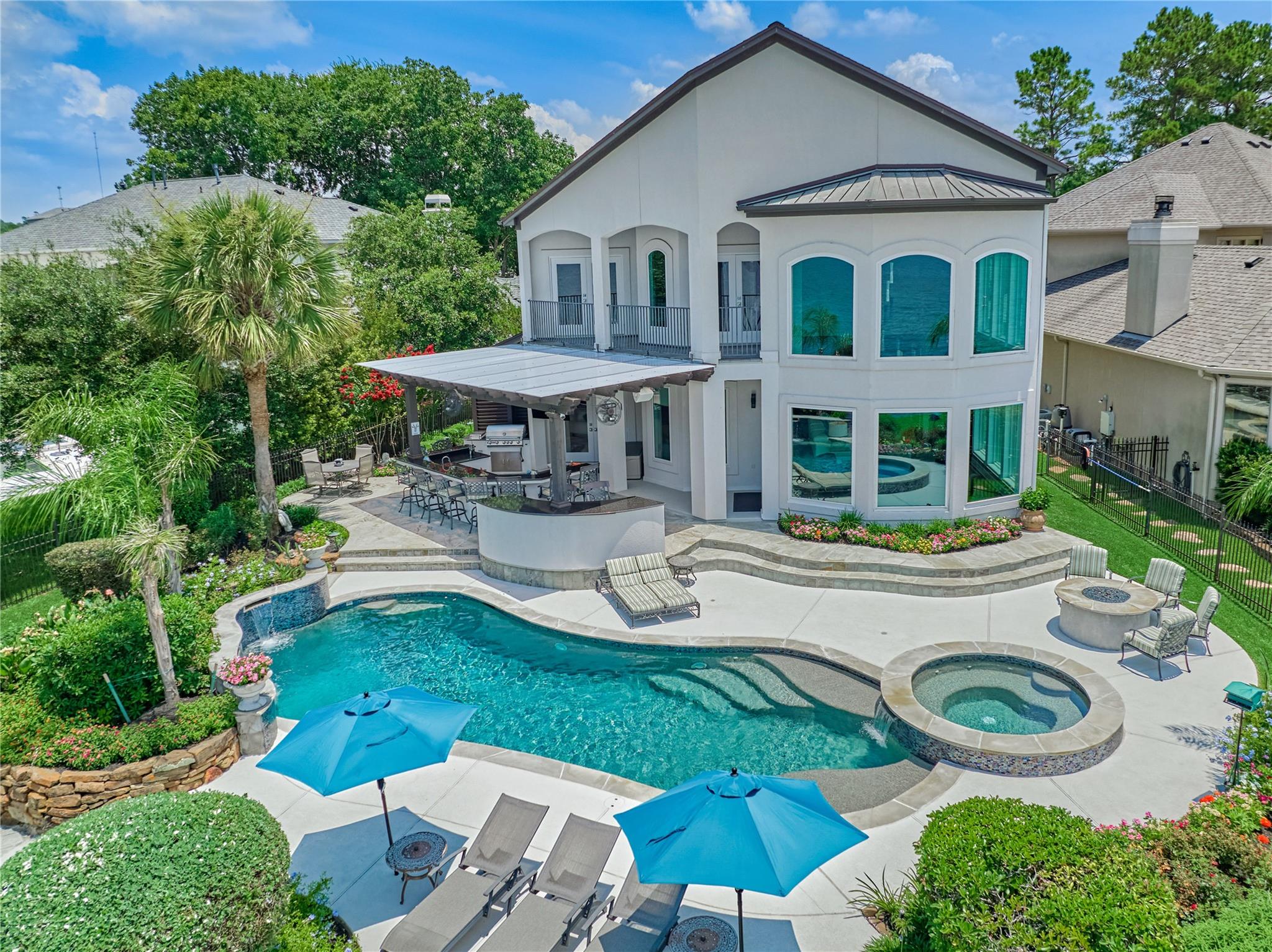 Two-story white home overlooking a landscaped pool and spa, with a covered outdoor kitchen and blue umbrellas.