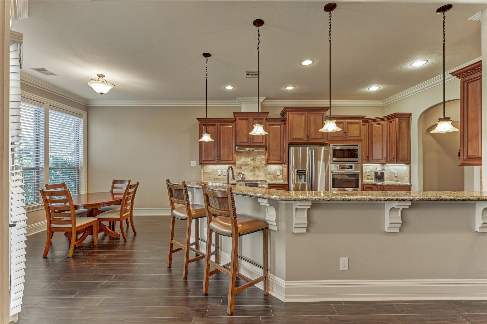Kitchen with granite island, bar stools, reddish wood cabinets, and dining table beside large windows.
