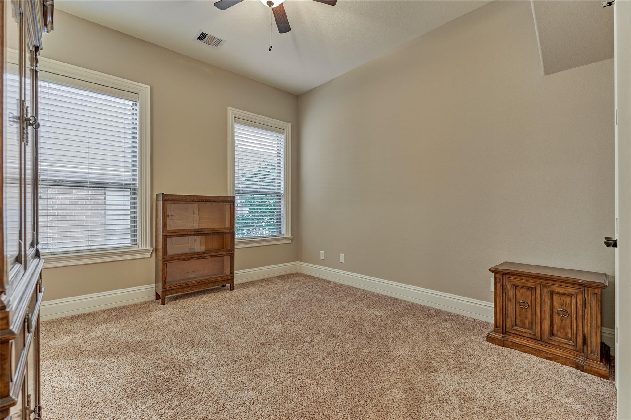 Carpeted bedroom with two shuttered windows, a ceiling fan, and wooden cabinets.