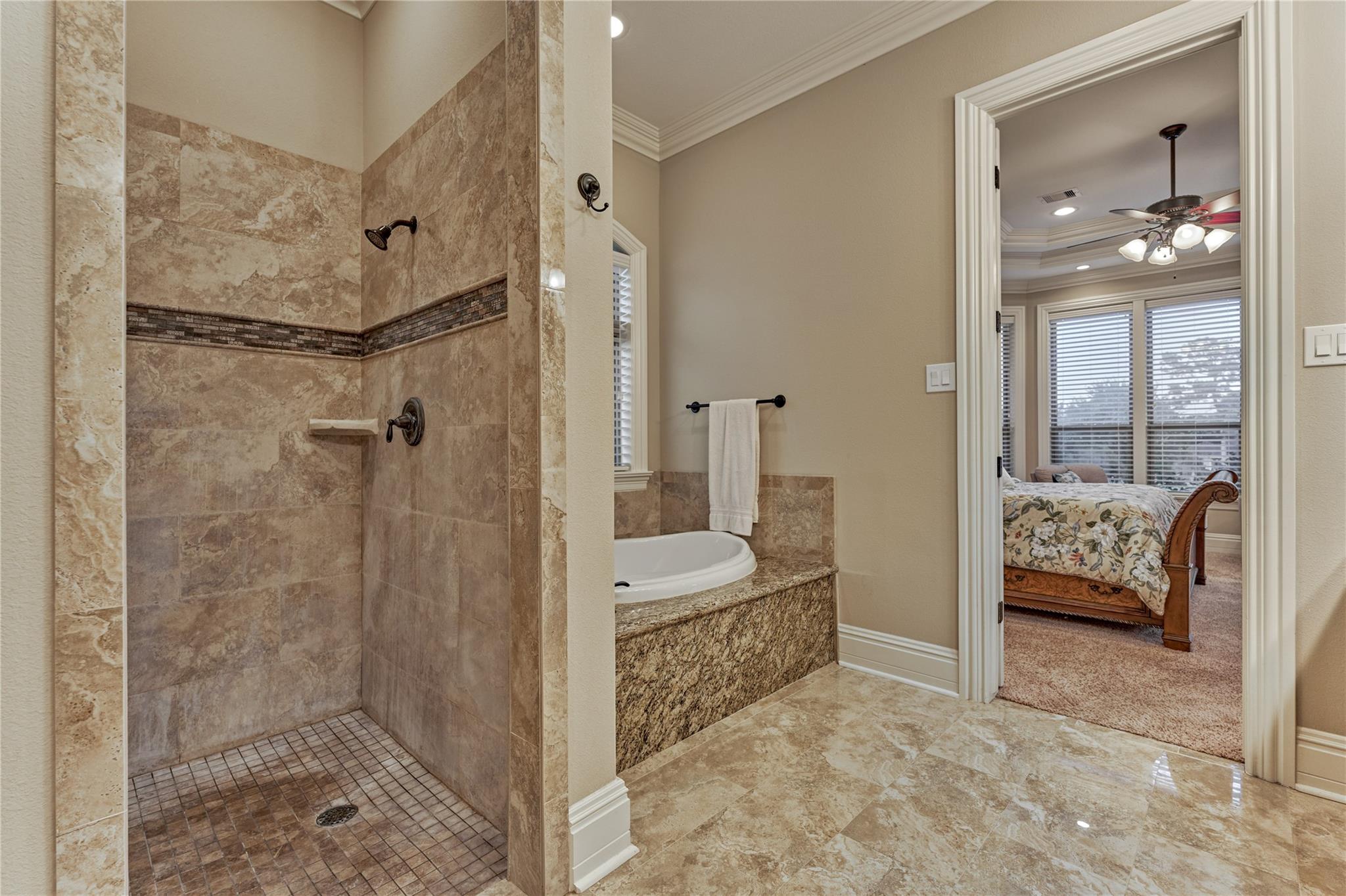 Bathroom with stone-tiled walk-in shower and soaking tub, opening onto a bedroom with a floral bedspread.