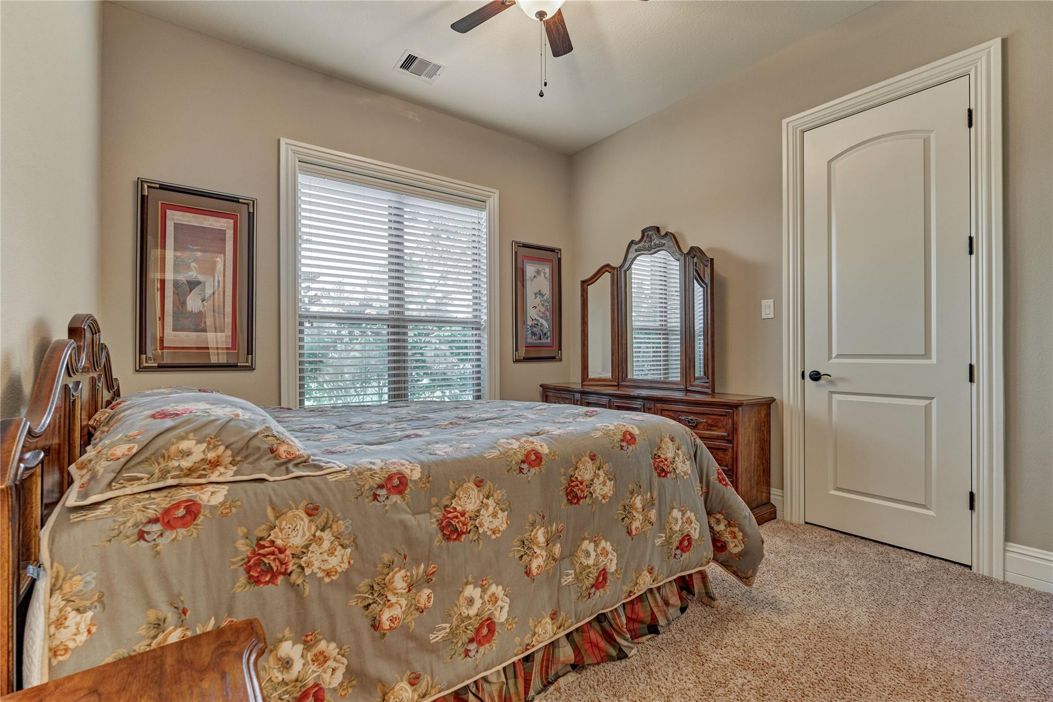 Furnished bedroom with a gray floral bedspread, carved wooden furniture, and a window with blinds.