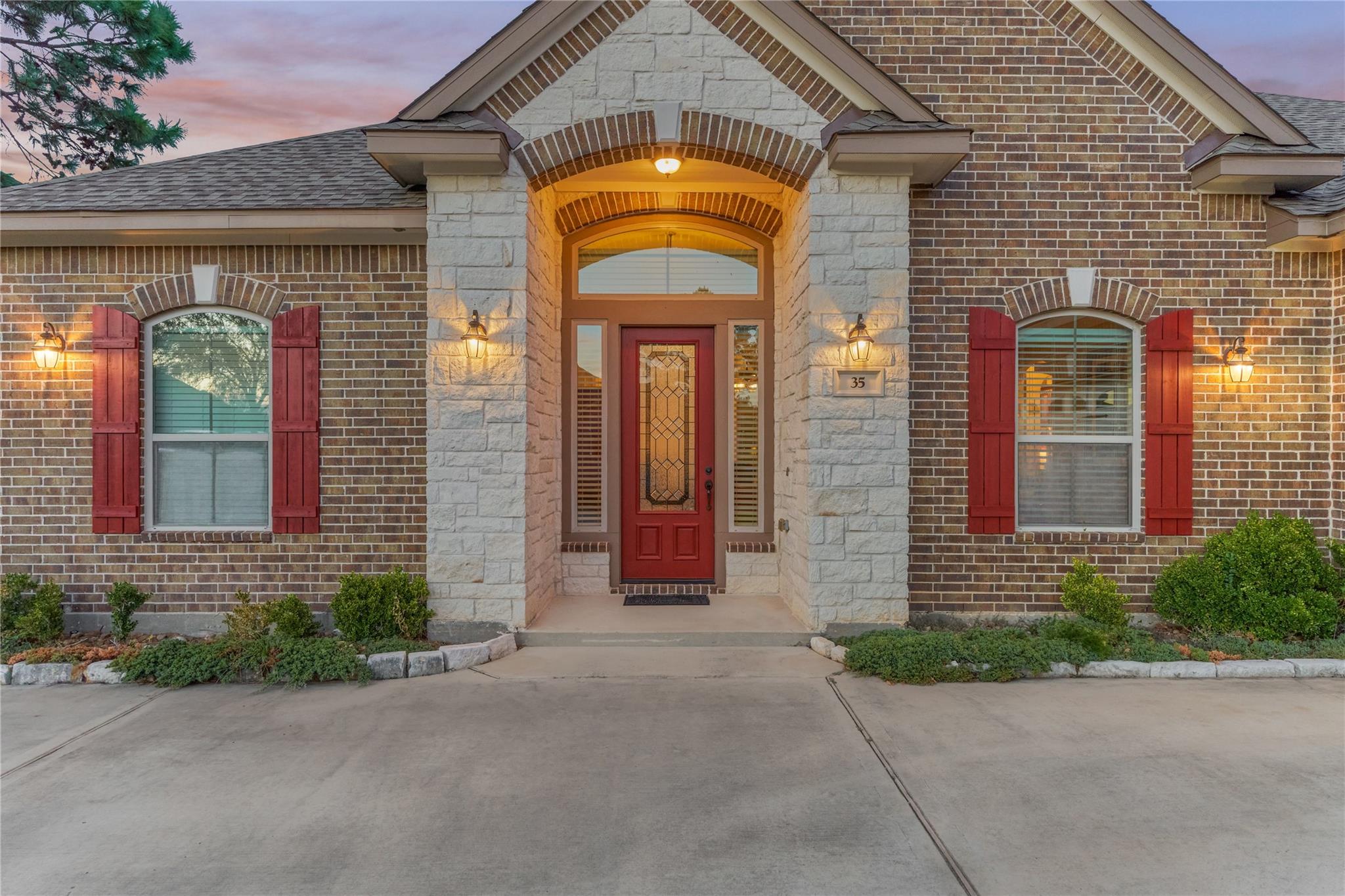 Brick home entrance with a stone arch, red front door and shutters at dusk.