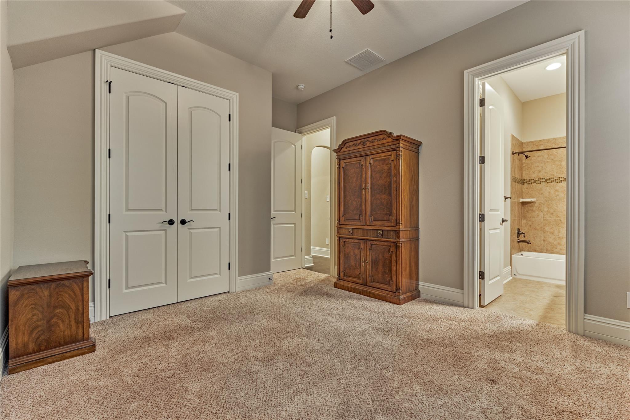 Bedroom with beige carpet and carved wooden armoire, with an adjoining tiled bathroom visible through the doorway.