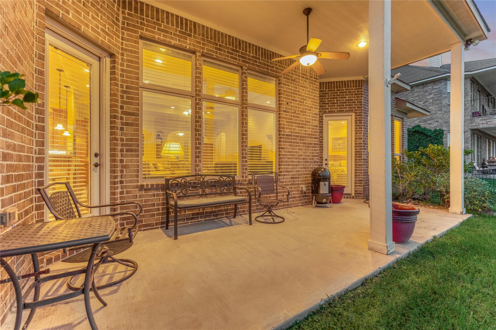 Covered brick patio with metal seating and a ceiling fan, warmly lit windows behind it.