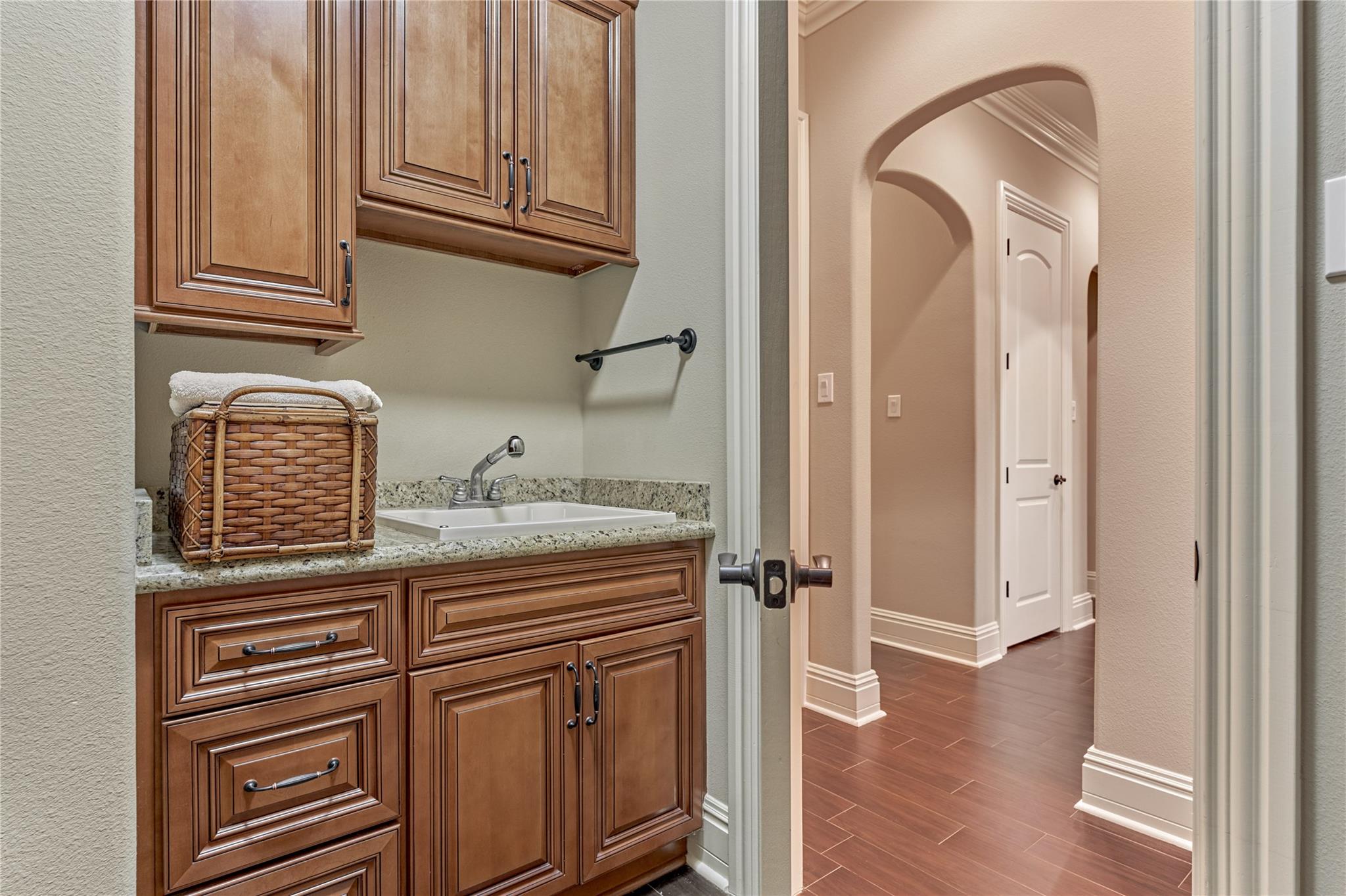 Utility room sink with dark wood cabinets, granite countertop, and a wicker basket of towels beside an arched hallway opening.