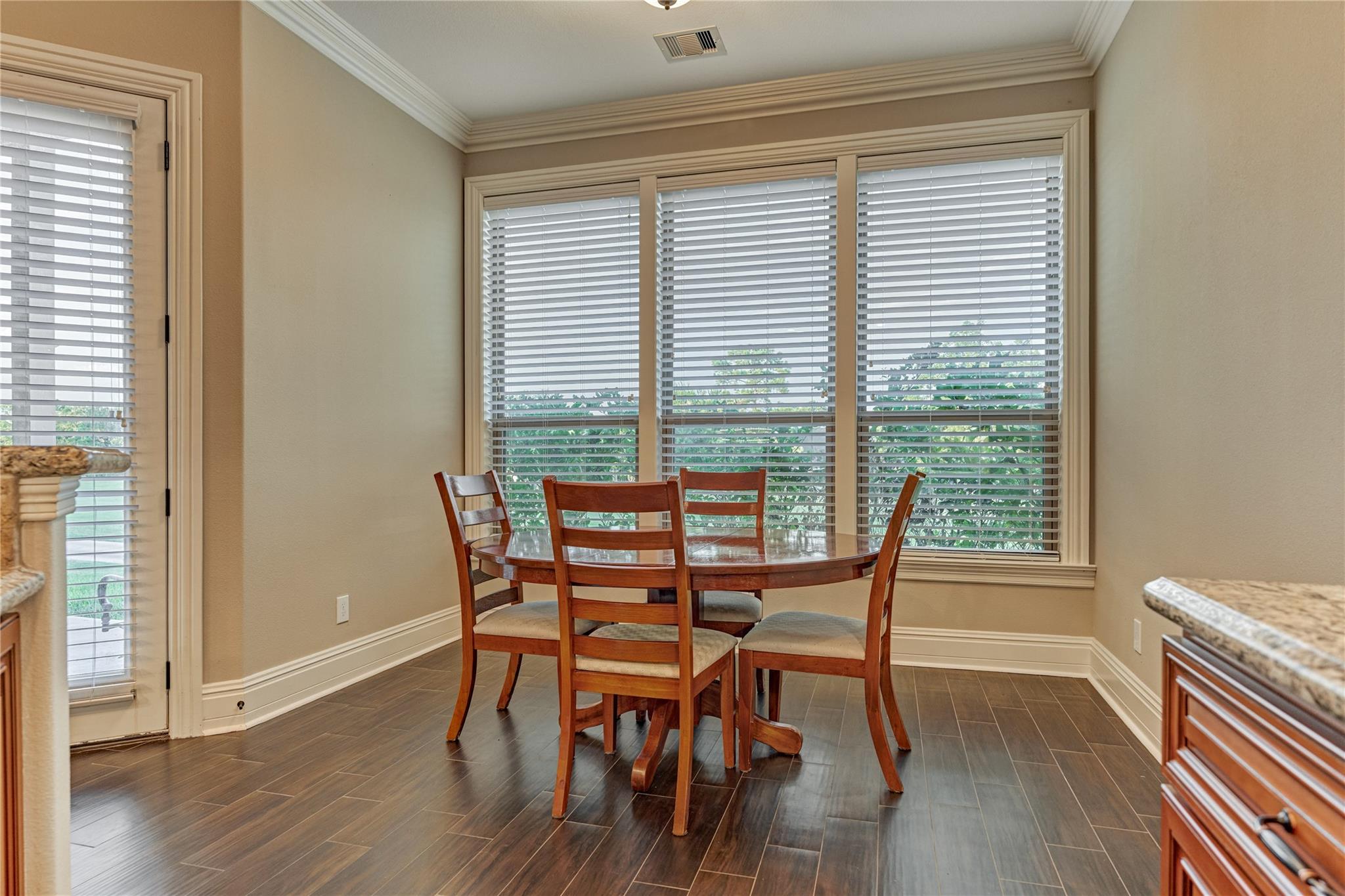 Dining area with a round wooden table, matching chairs, and large windows with white blinds.