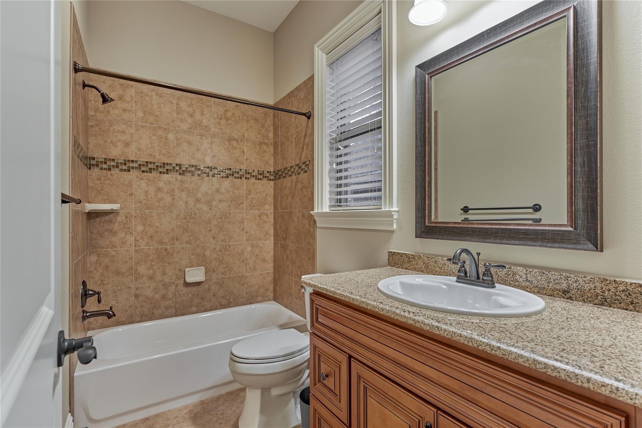 Bathroom with a tan tiled tub and shower, wood vanity, granite countertop, and window with blinds.