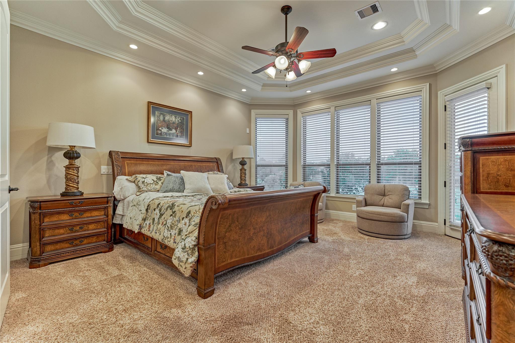 Bedroom with a carved wooden bed, floral bedding, tray ceiling, and wide windows with blinds.