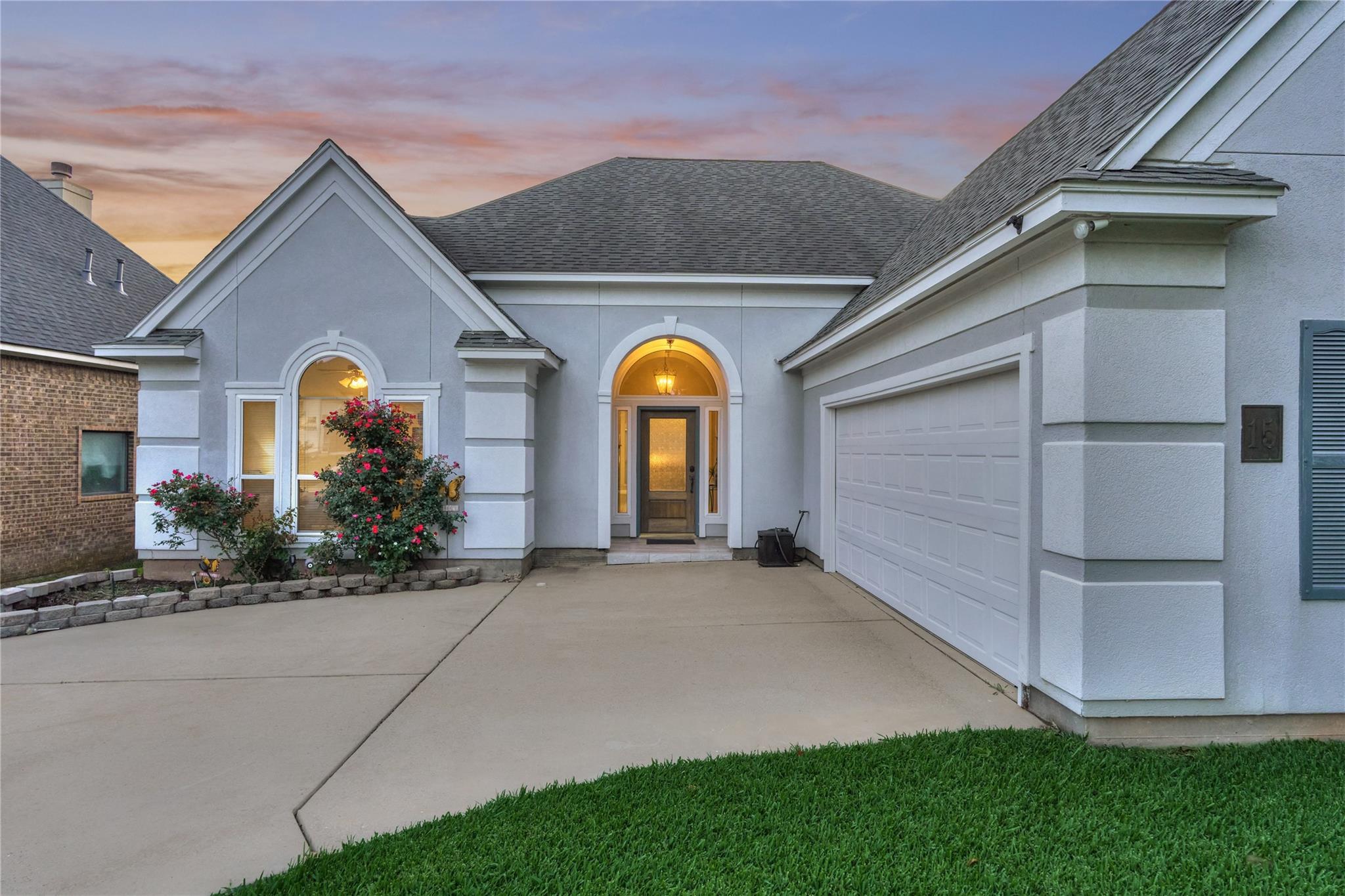 Gray stucco home with an arched entry, two-car garage, and flowering shrubs at dusk.