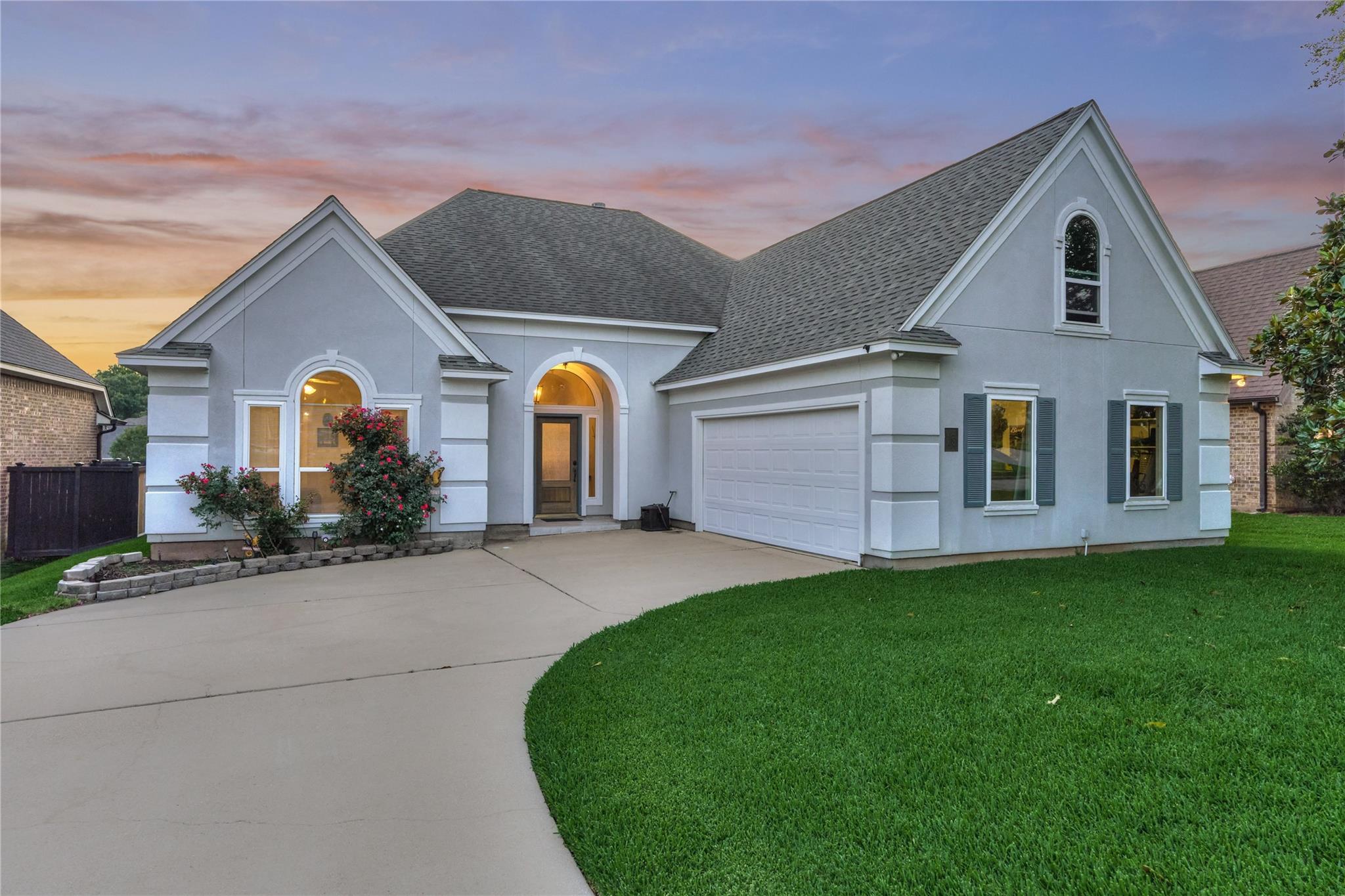 Light gray home with steep gables, arched entry, double garage, and curved driveway at dusk.