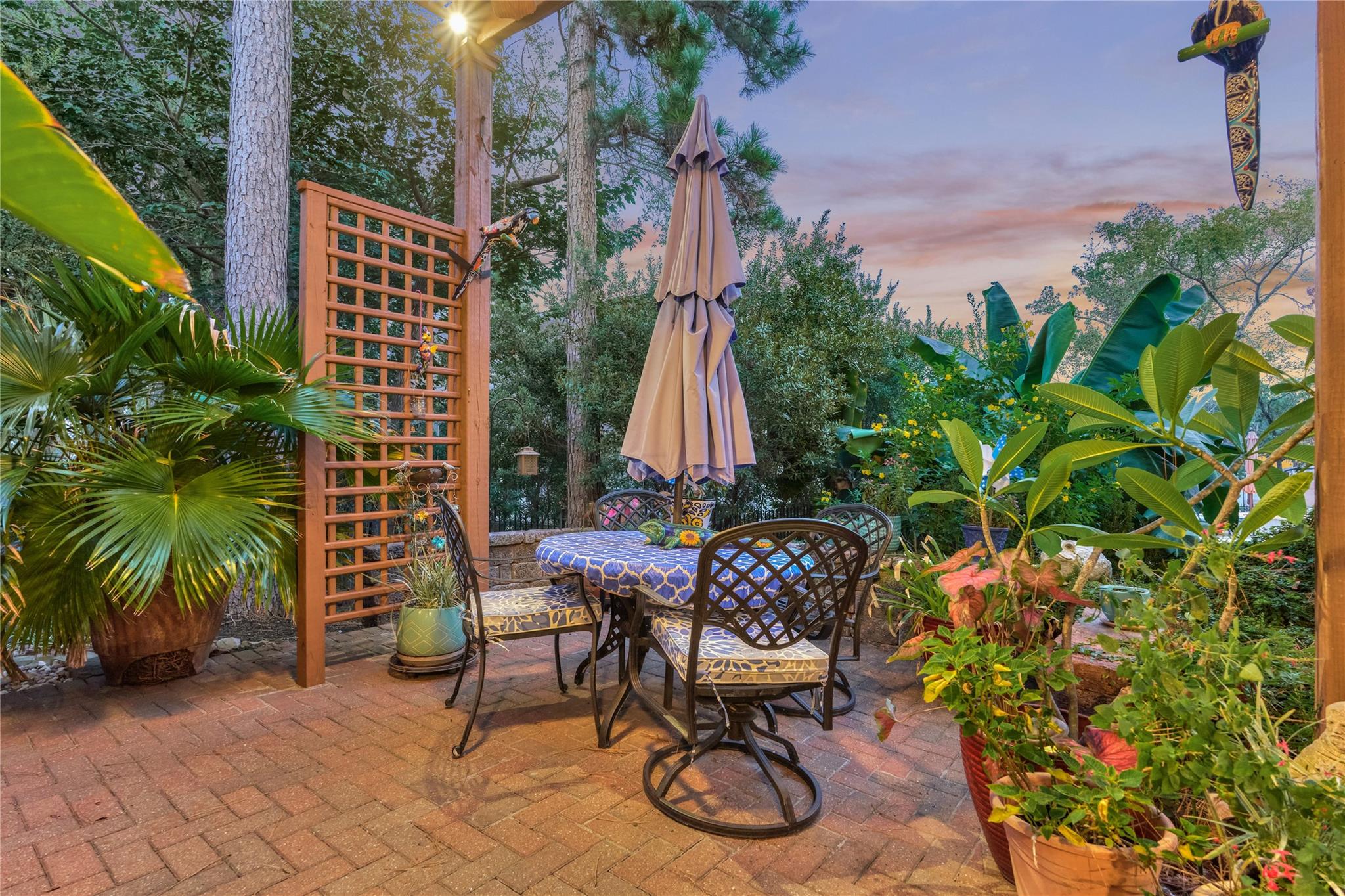 Furnished brick patio with a dining set, closed umbrella, wooden trellis, and lush potted plants.