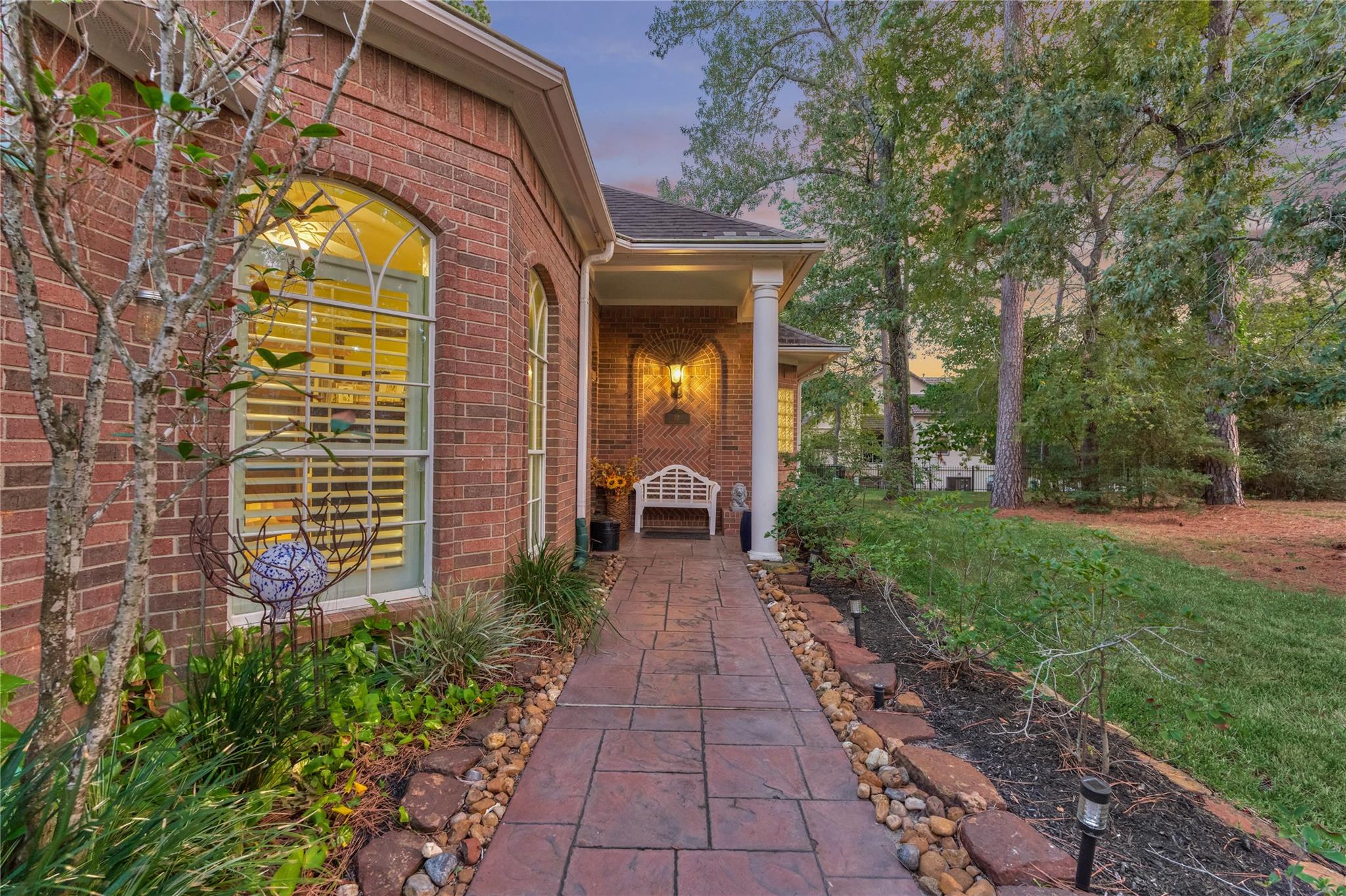 Brick home entrance with a covered porch, white bench, and stamped-concrete walkway at dusk.