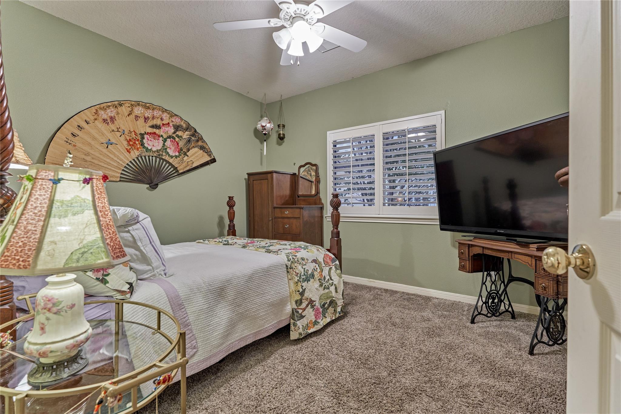Bedroom with floral bedding, sage-green walls, plantation shutters, and a large television.