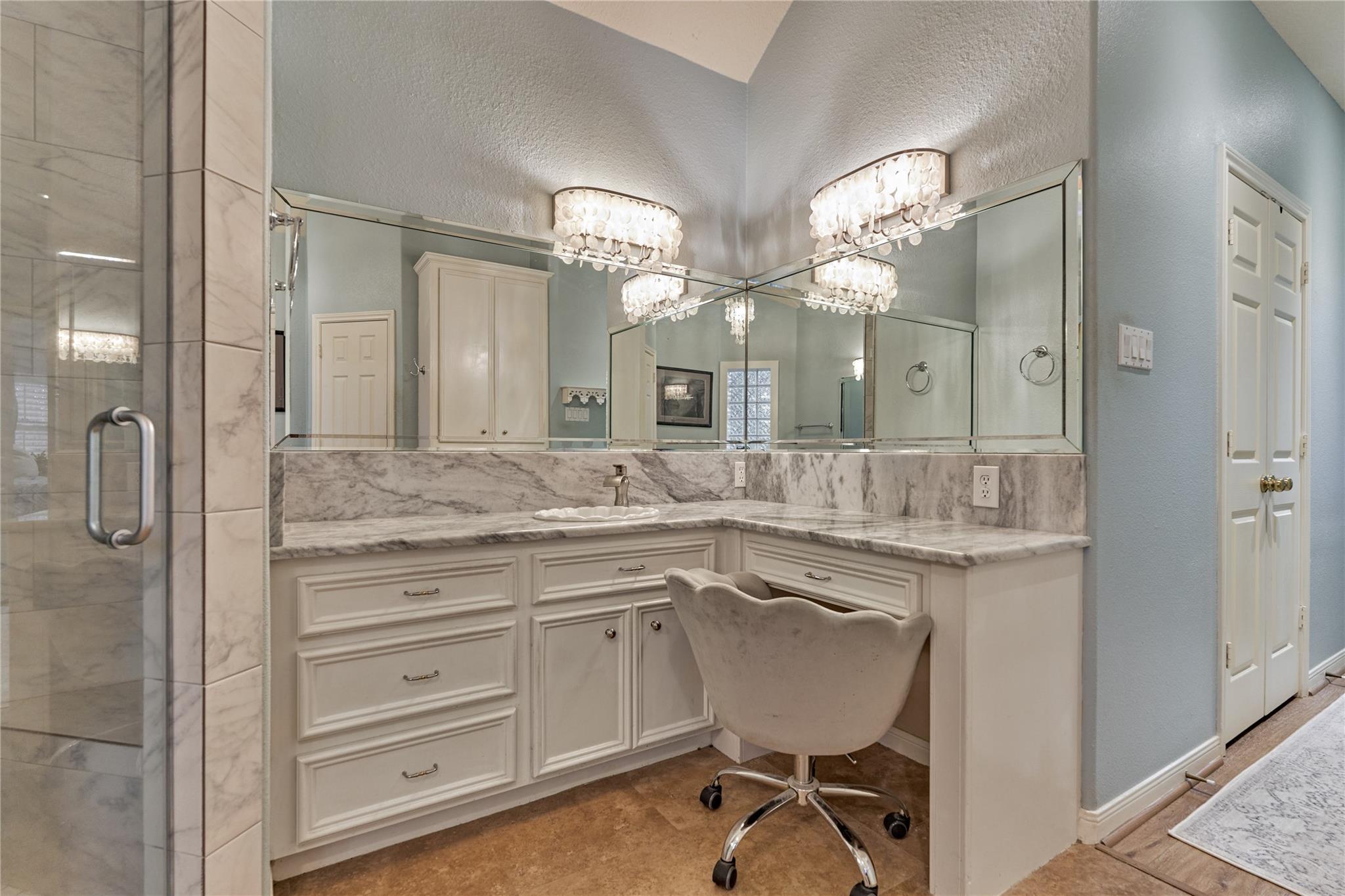 Bathroom vanity with a marble countertop, mirrored wall, glass shower, and cushioned swivel chair.