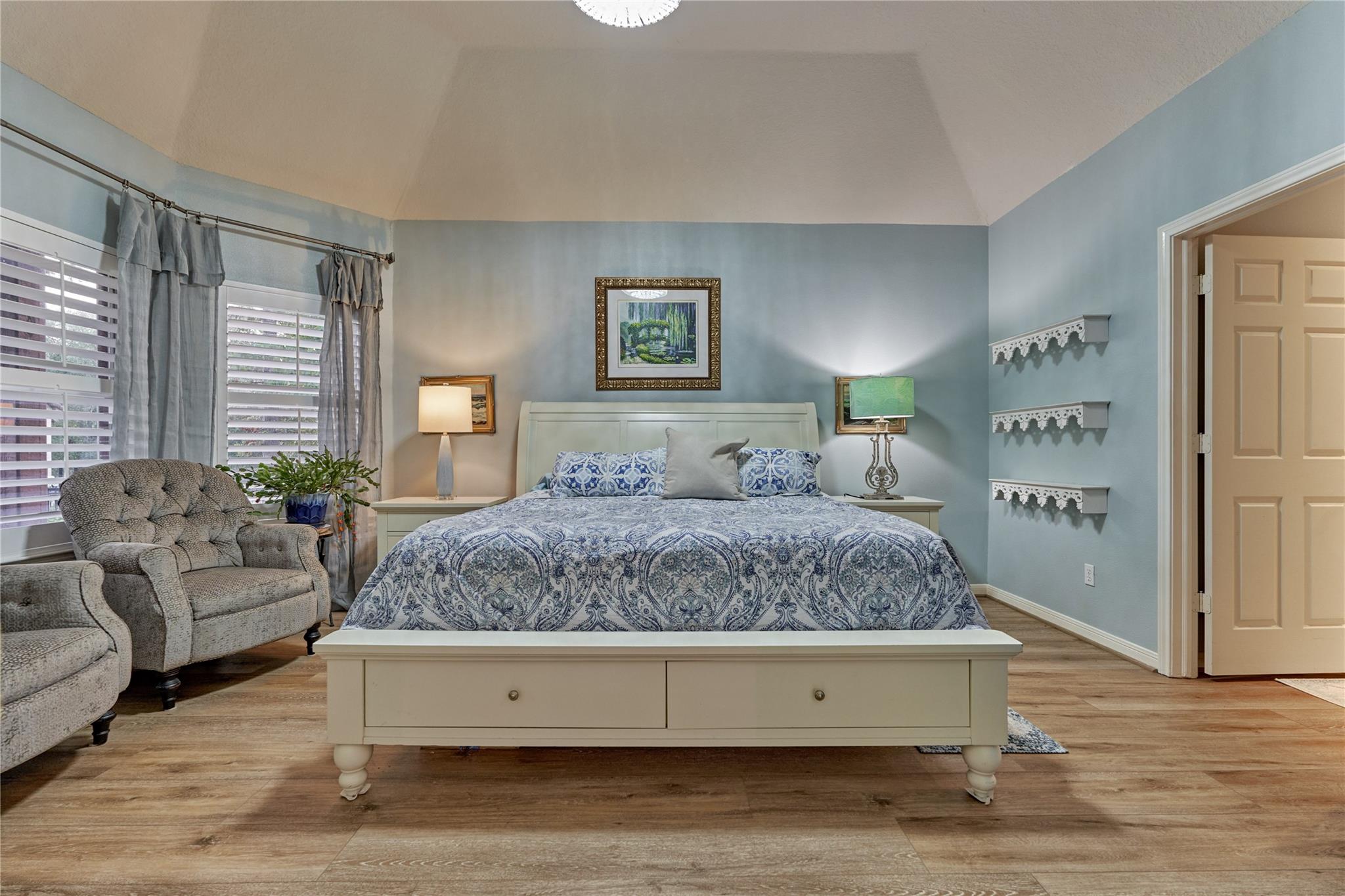 Bedroom with a white bed, blue patterned bedding, pale blue walls, and a chair by shuttered windows.