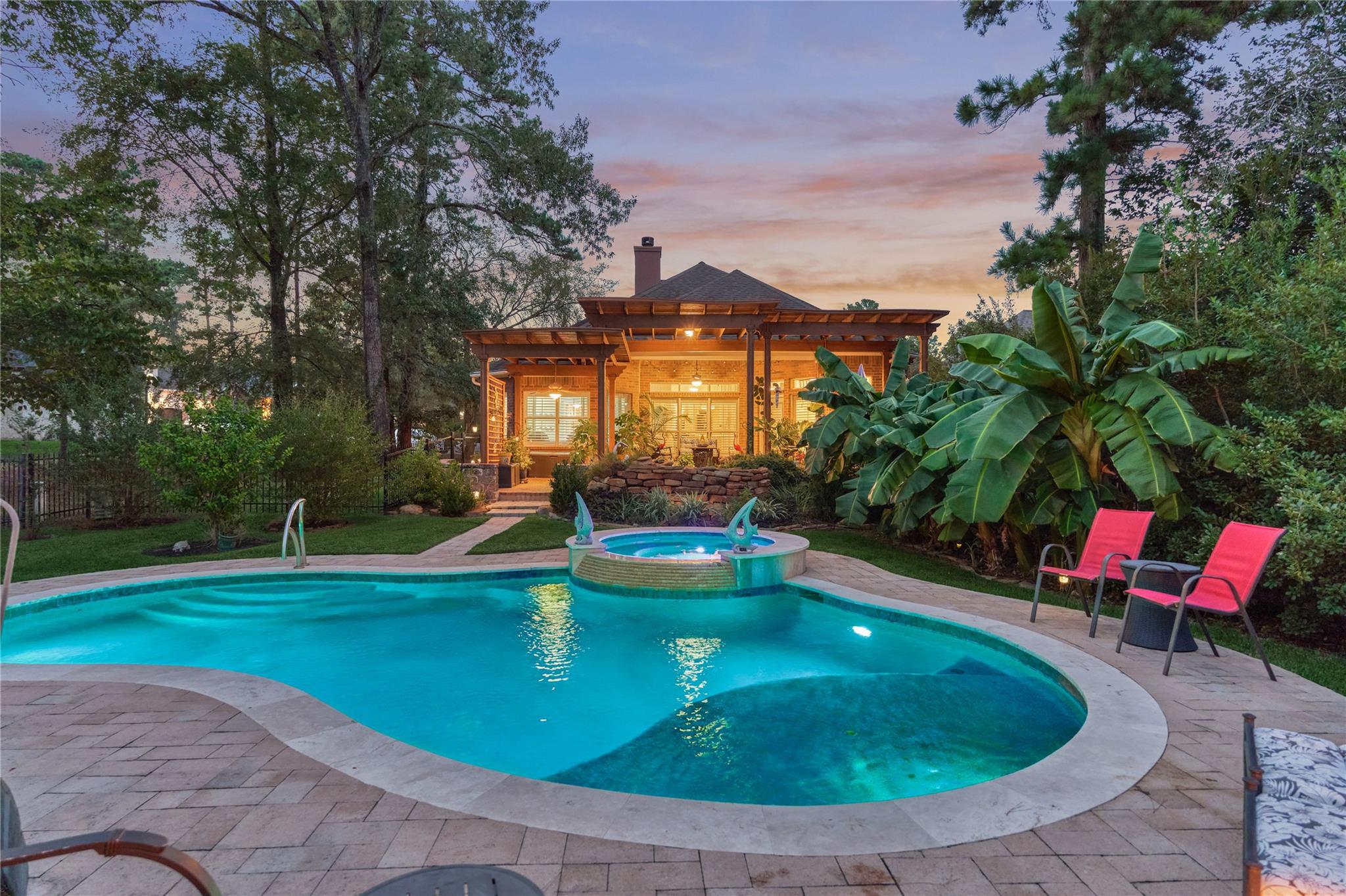 Backyard pool and raised spa beside a warmly lit covered porch, surrounded by tropical plants and patio seating.