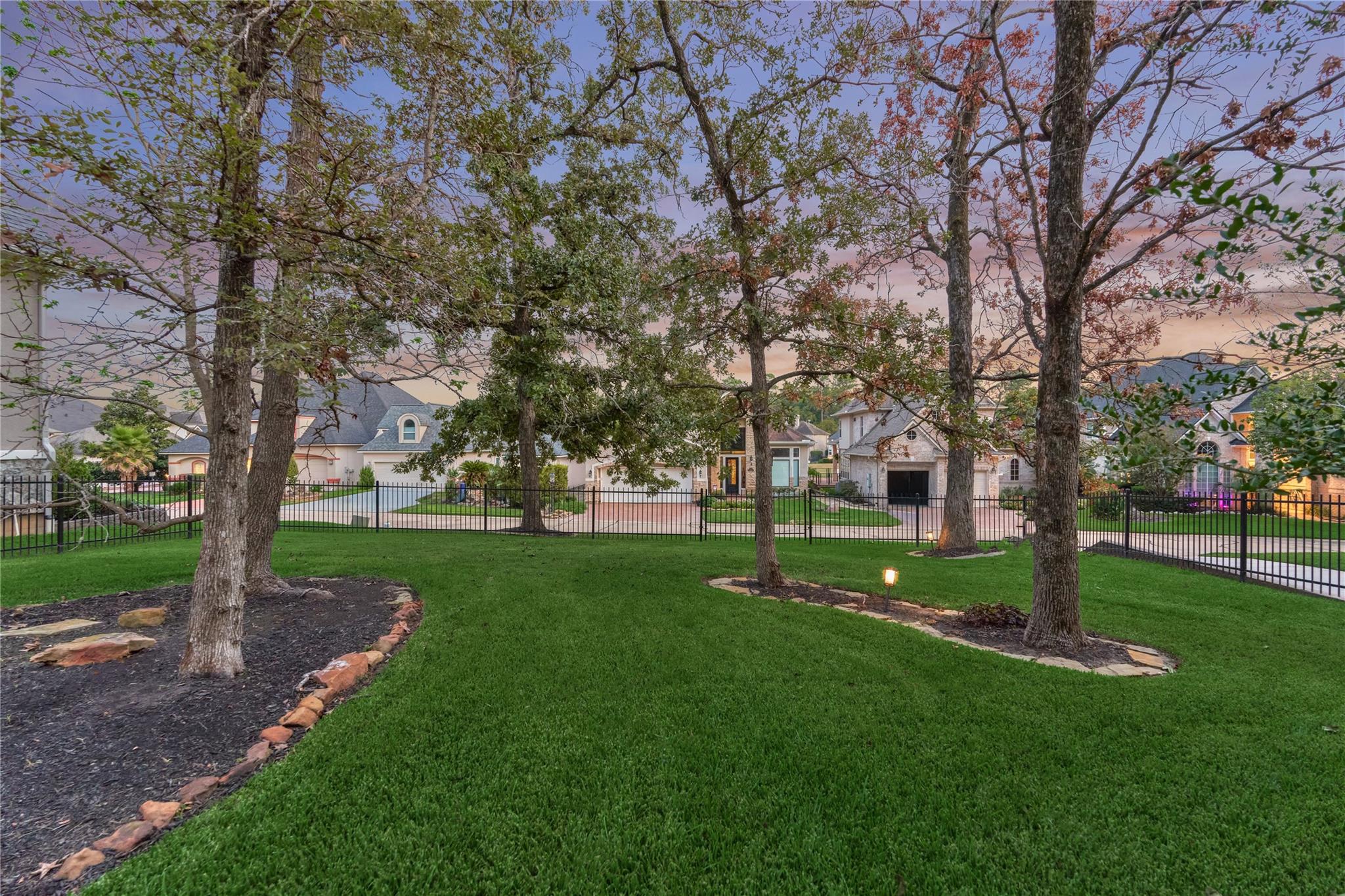 Tree-shaded lawn with landscaped beds and a black metal fence at dusk.