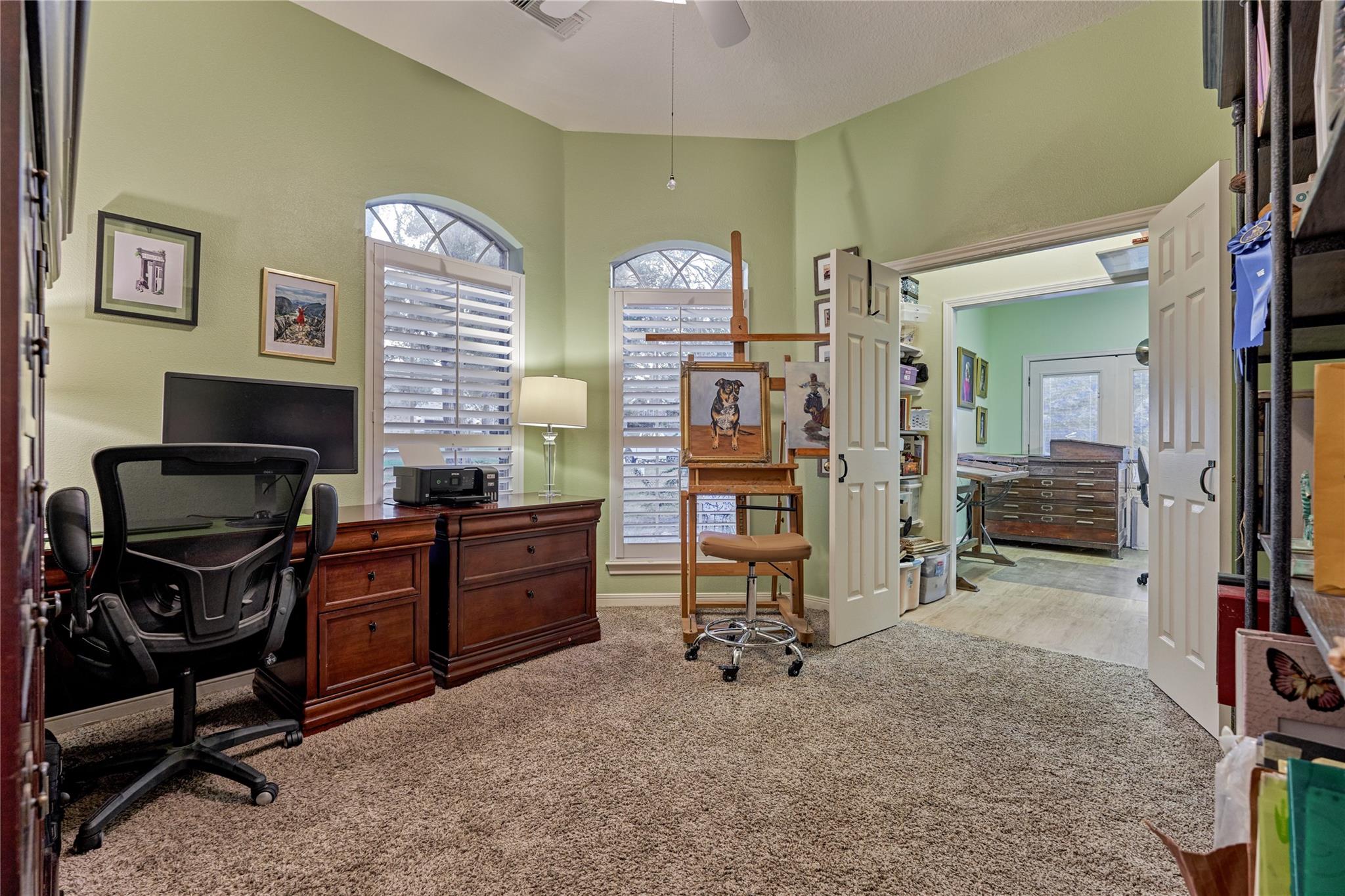 Home office with arched shuttered windows, a desk, and an easel holding a dog portrait; open doors lead to another workspace.