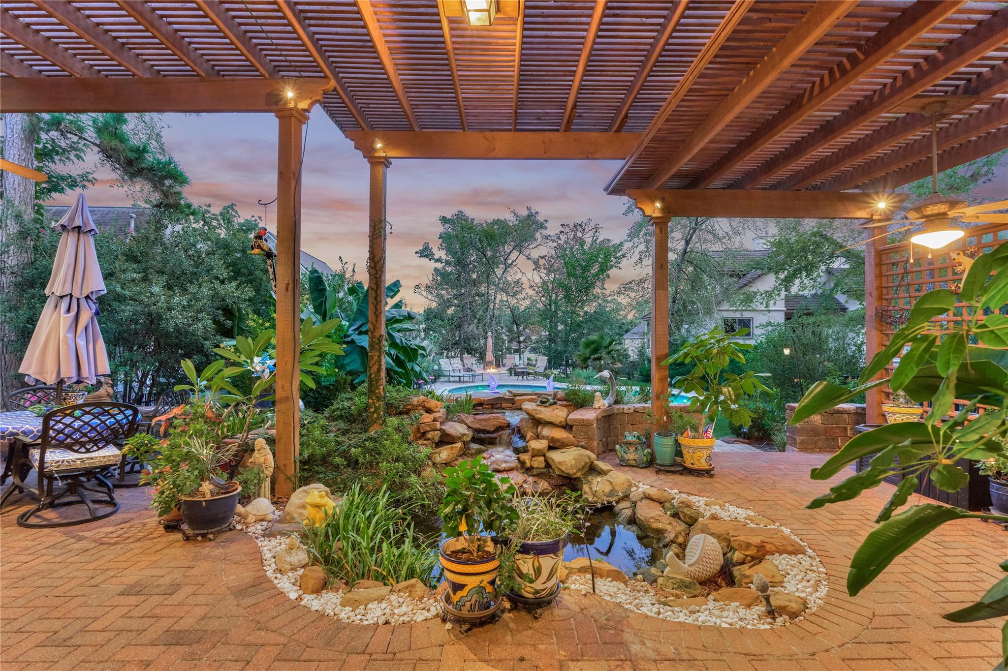 Backyard patio beneath a wooden pergola, with a rock-lined pond, garden plants, and pool beyond.