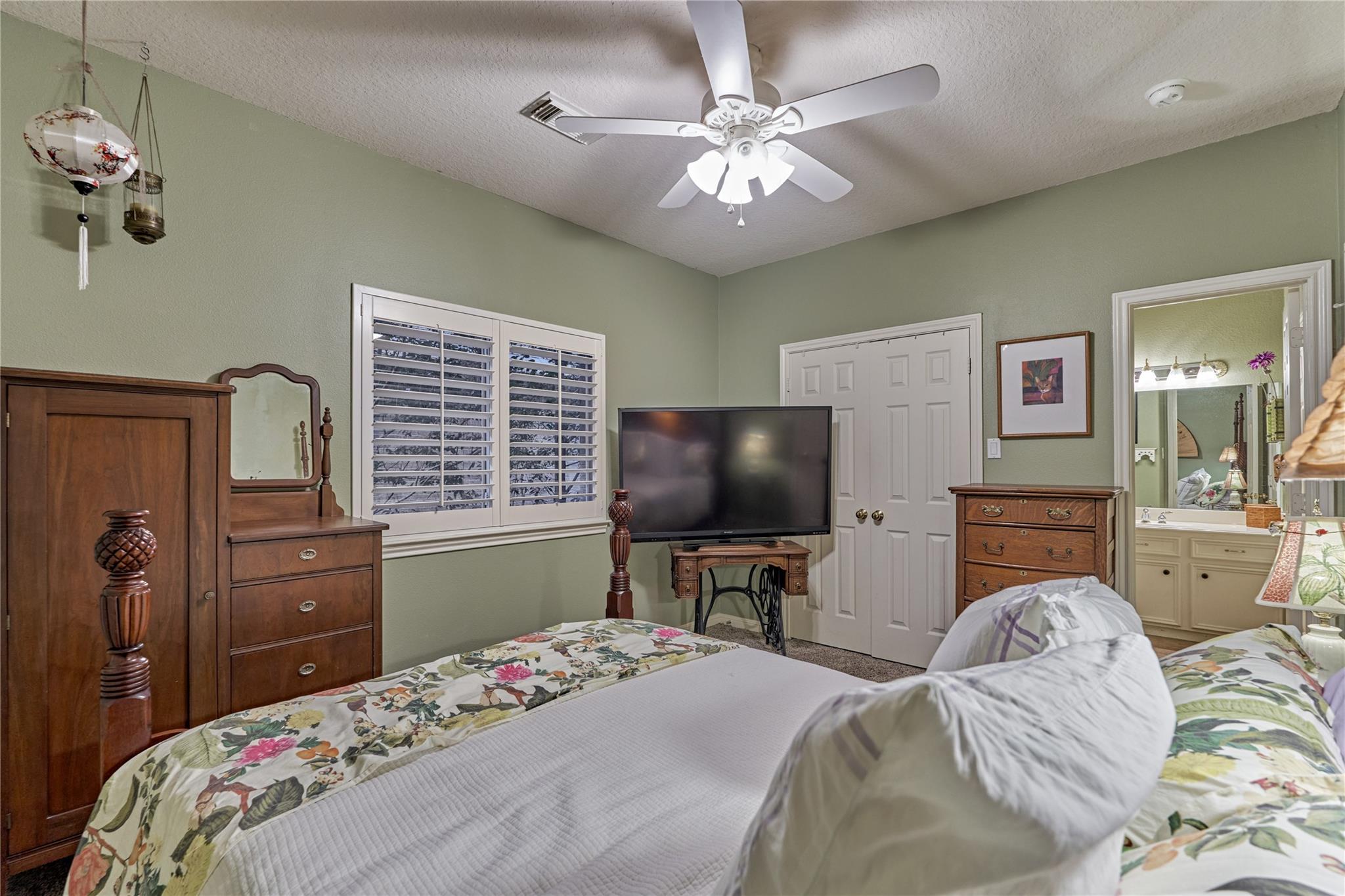 Bedroom with floral bedding, wooden dressers, plantation shutters, and a ceiling fan.