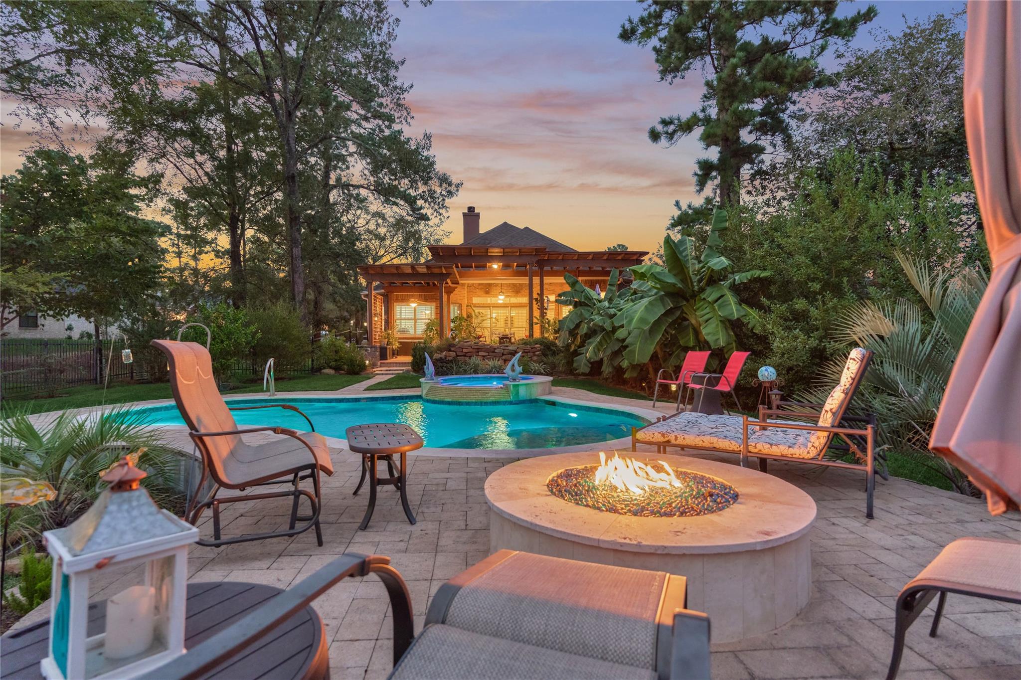 Backyard pool and raised spa beside a paved patio with seating and a glowing fire pit at dusk.