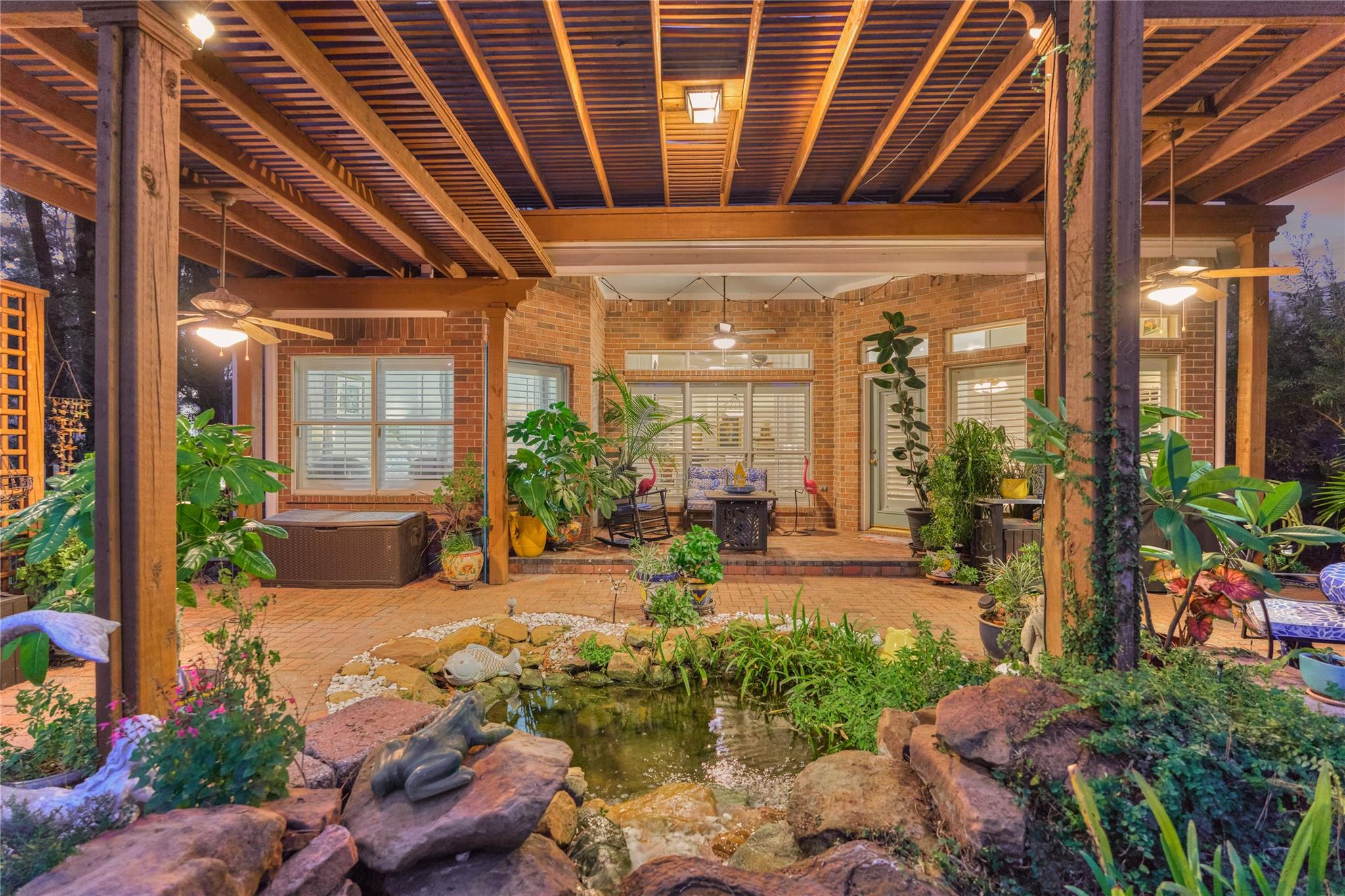 Covered brick patio with timber pergola, abundant plants, and a rock-edged garden pond.