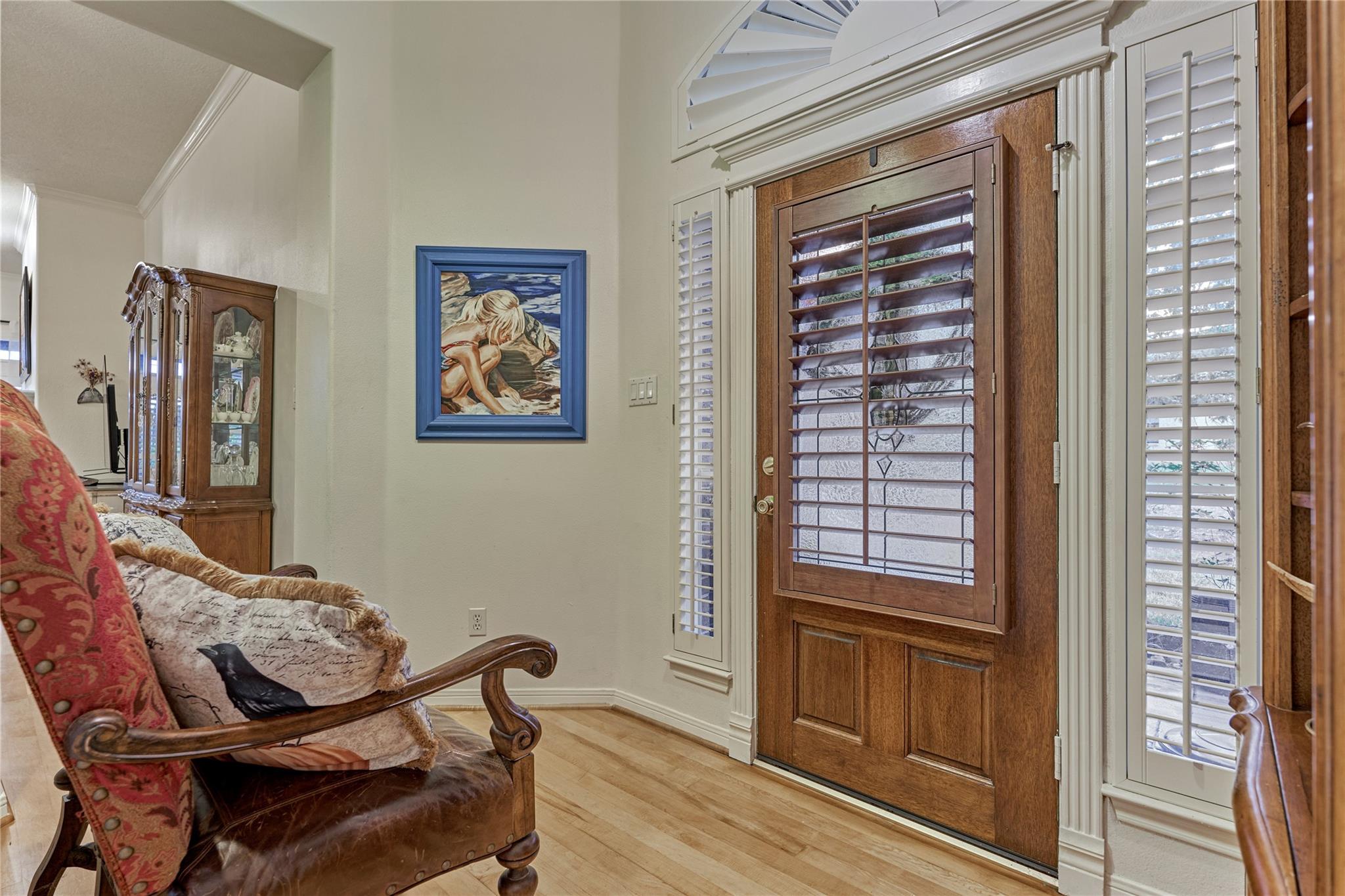 Foyer with a shuttered wood front door, patterned armchair, display cabinet, and light wood flooring.