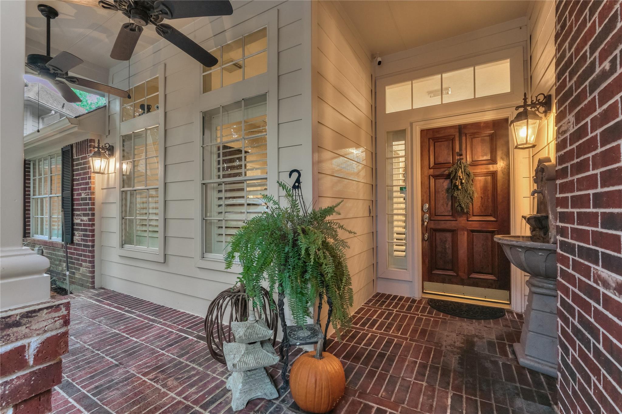 Covered brick front porch with a dark wooden door, glowing lanterns, potted ferns, and a small fountain.