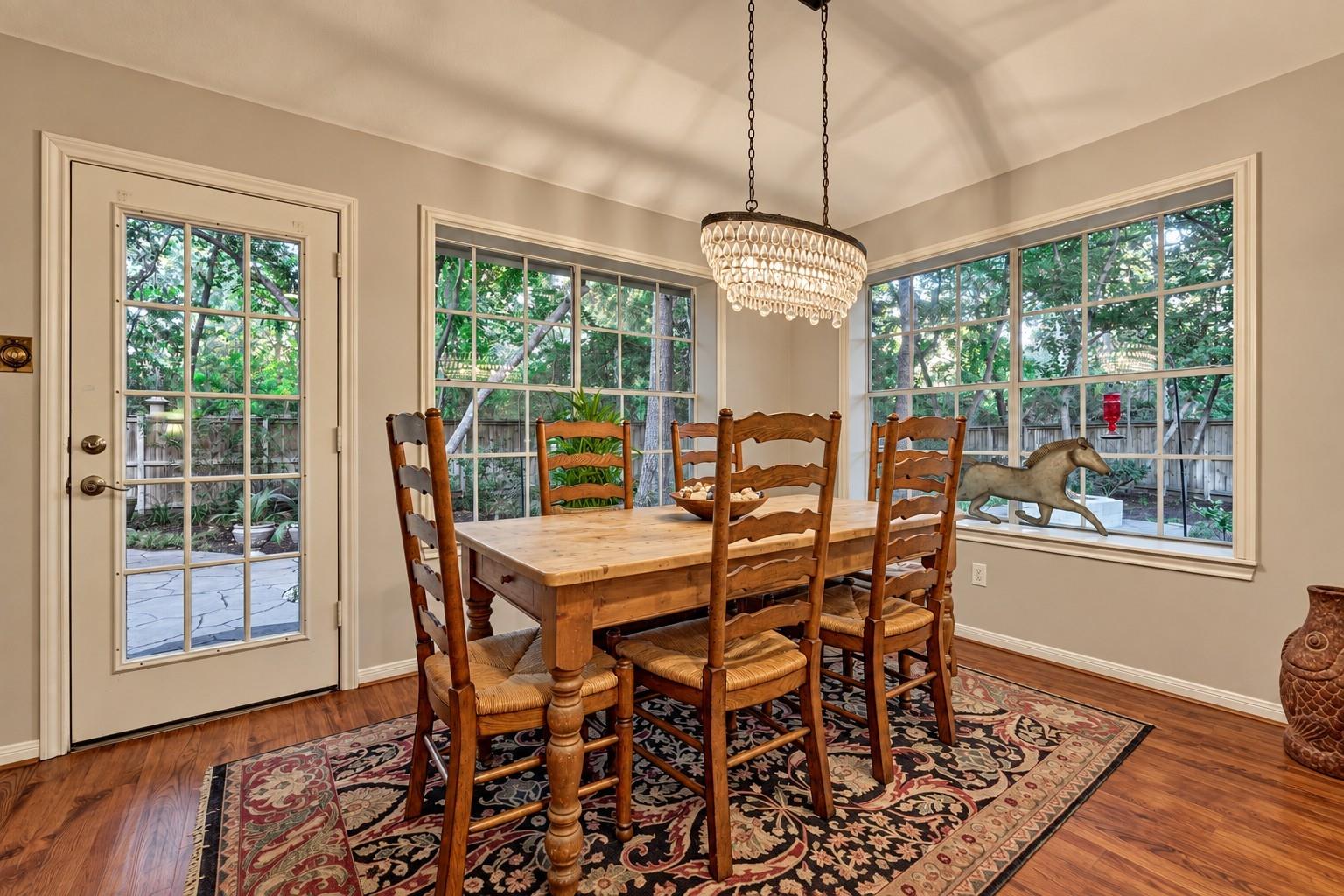 Dining room with a wooden table, crystal chandelier, and large windows overlooking a leafy yard.