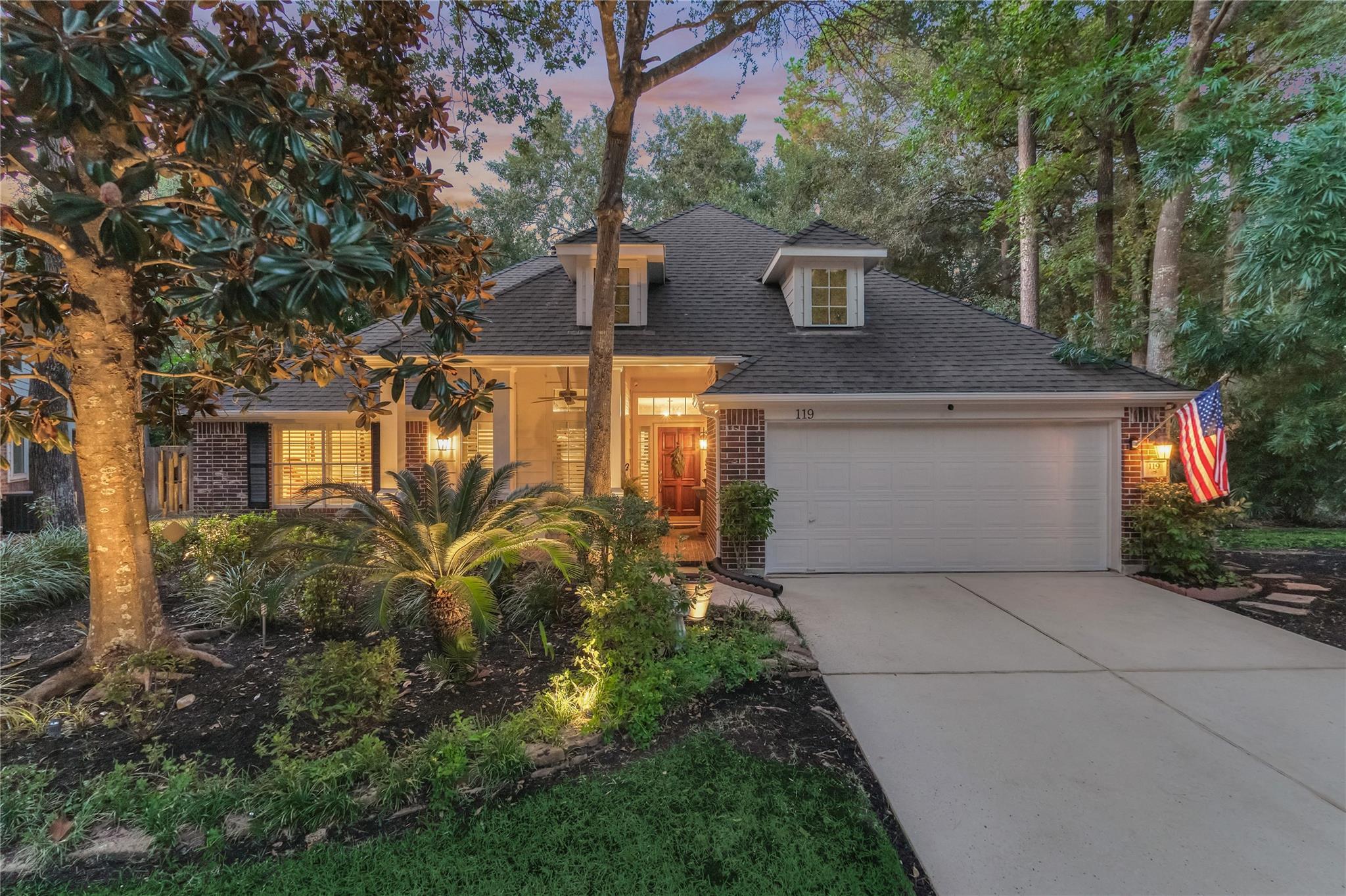 Brick home at dusk with a lit covered entry, landscaped garden, and American flag beside the garage.