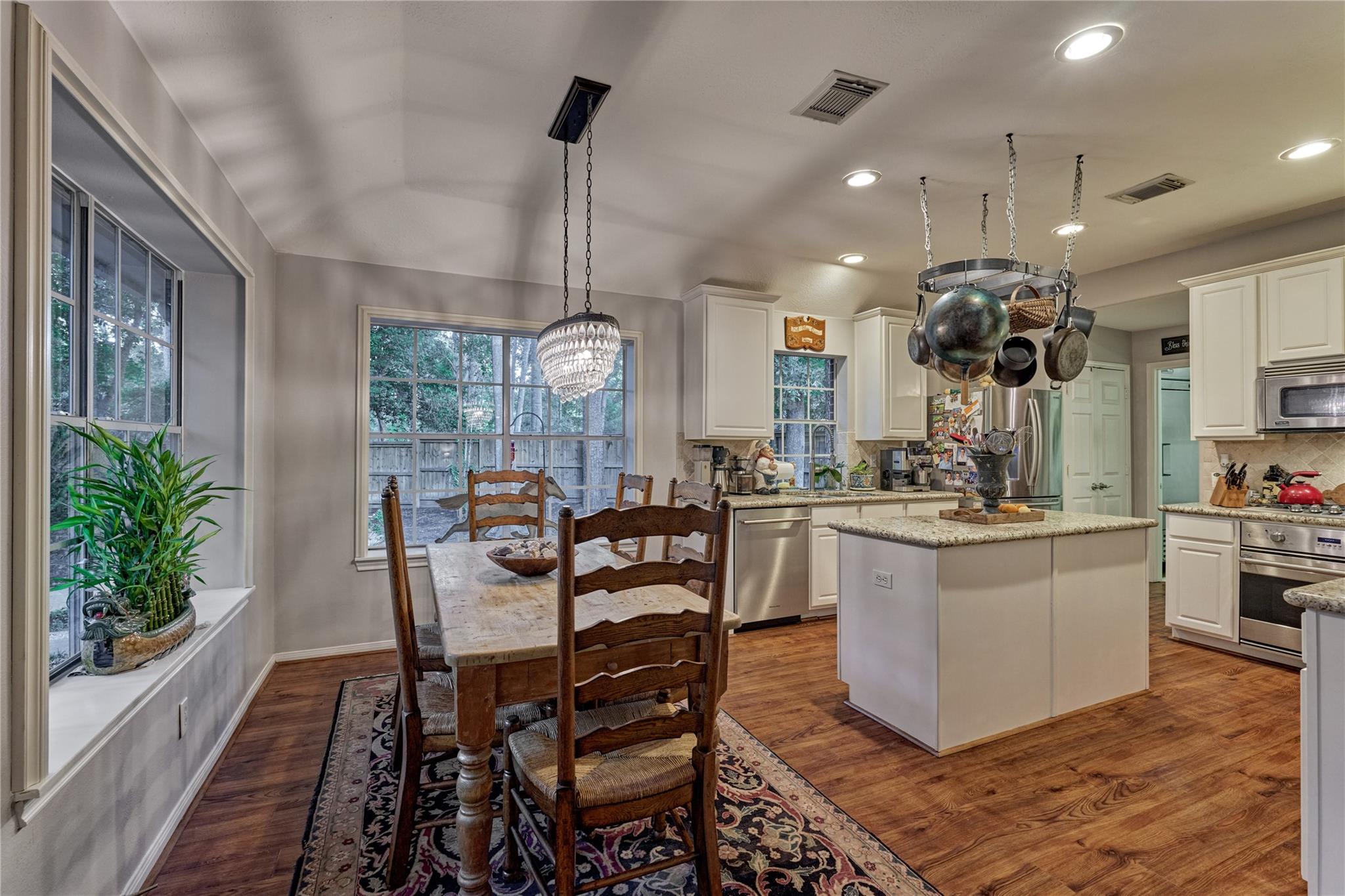 Kitchen and dining area with a granite island, hanging cookware, and a rustic table by large windows.