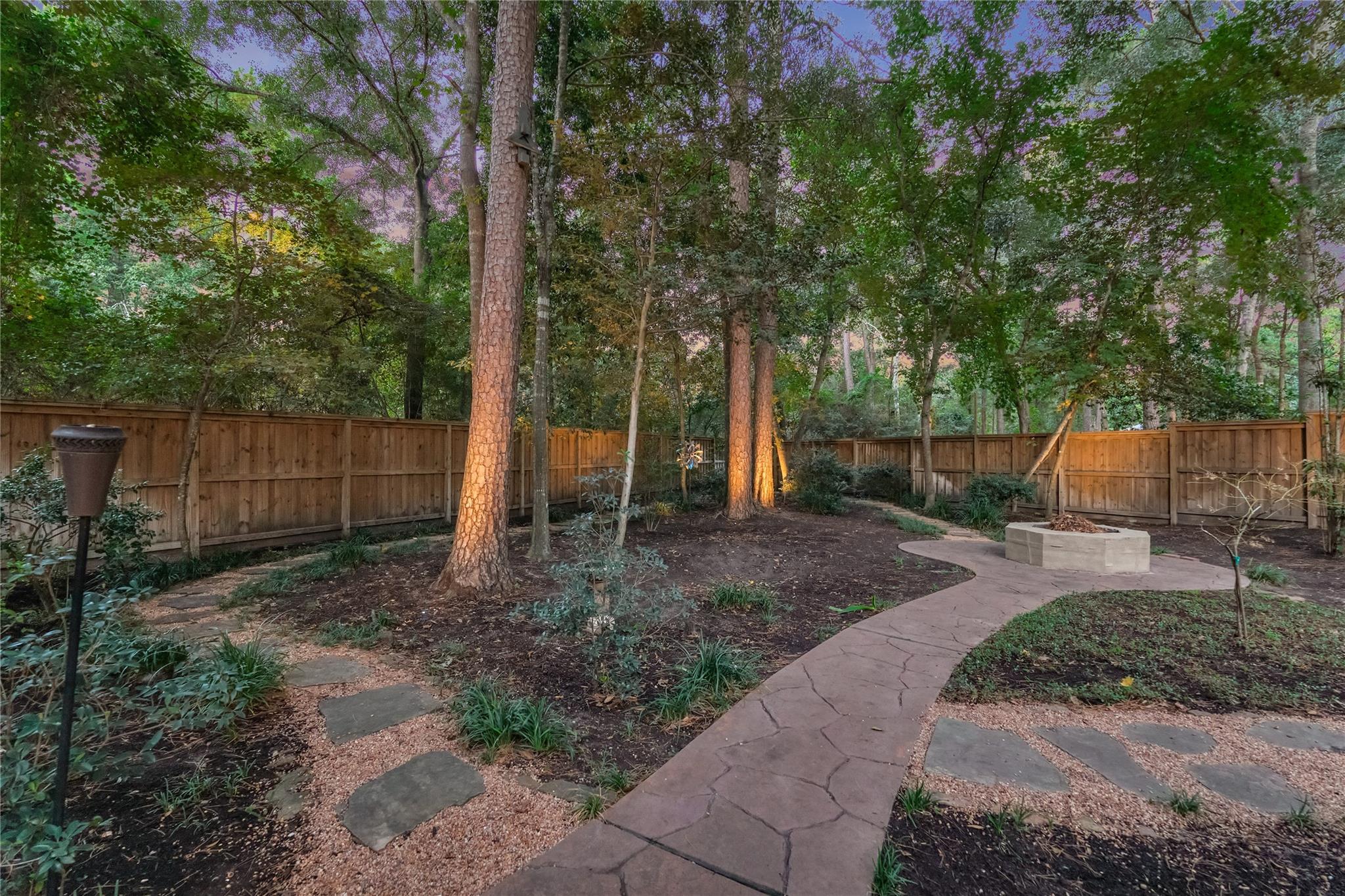 Wooded backyard with a curving path leading to a raised fire pit, enclosed by a wooden fence.